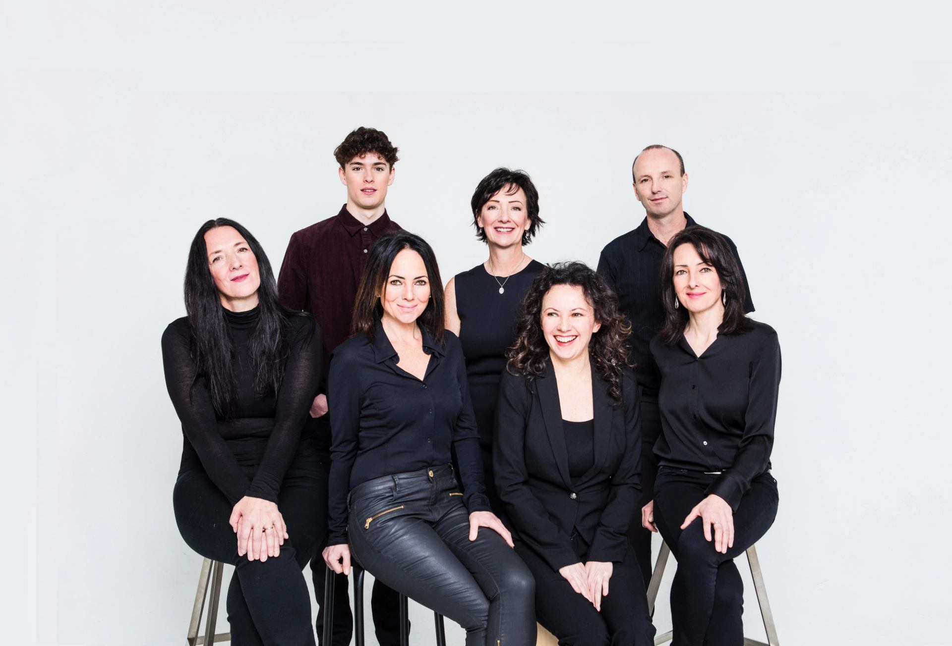 Group portrait of eight people in black clothing sitting and standing against white background.