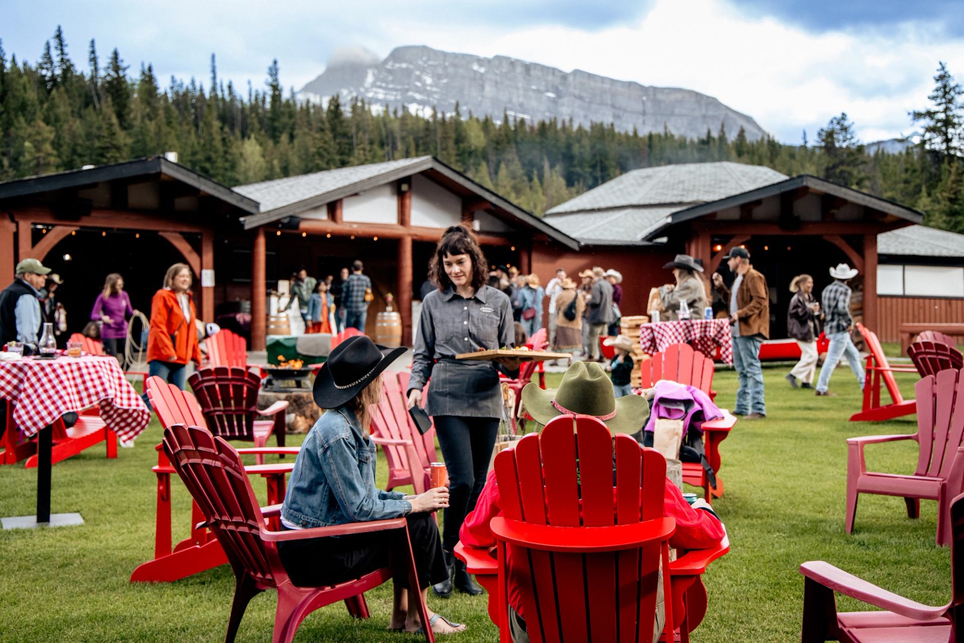 An outdoor event with people in red chairs on a lawn, backed by rustic buildings and mountains.