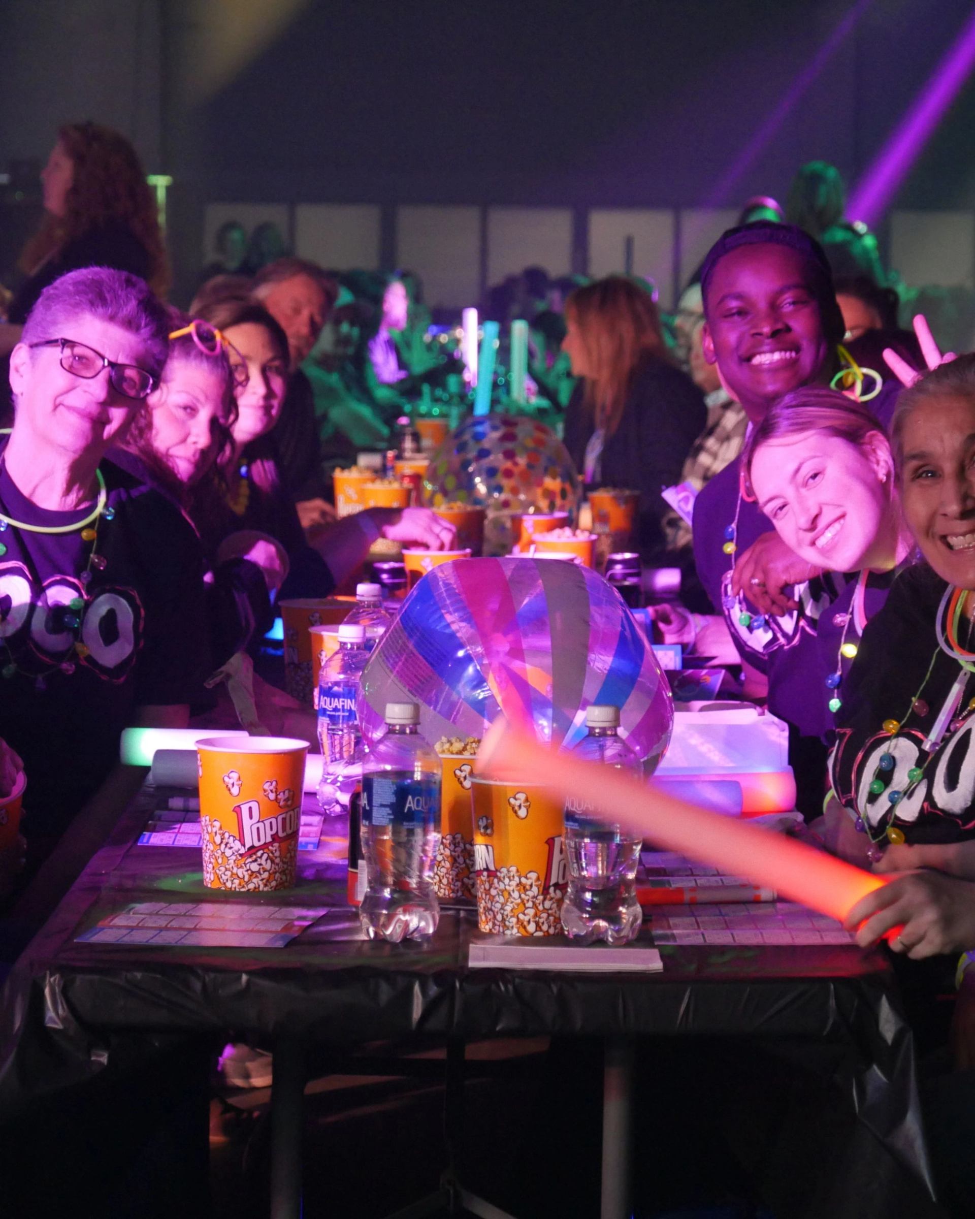 Friends at a neon-lit party table enjoying snacks and drinks together.