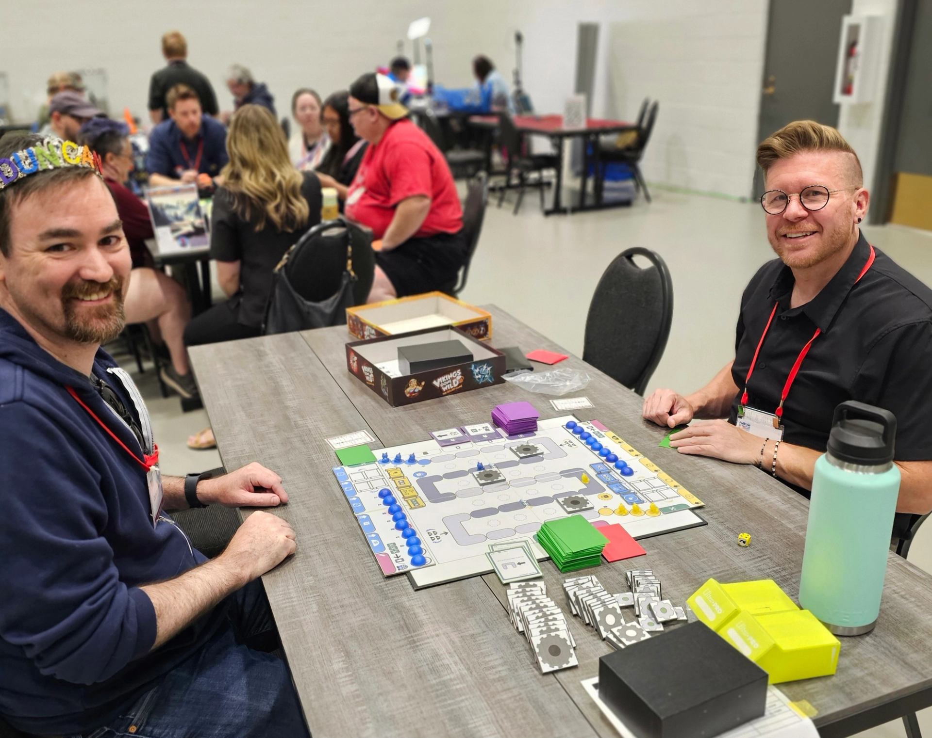 People sit at tables in a hall, playing a colourful board game together.