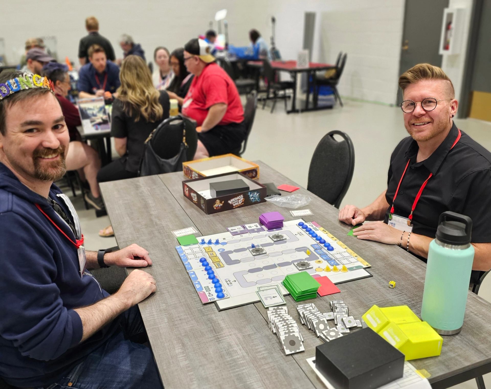 People sit at tables in a hall, playing a colourful board game together.