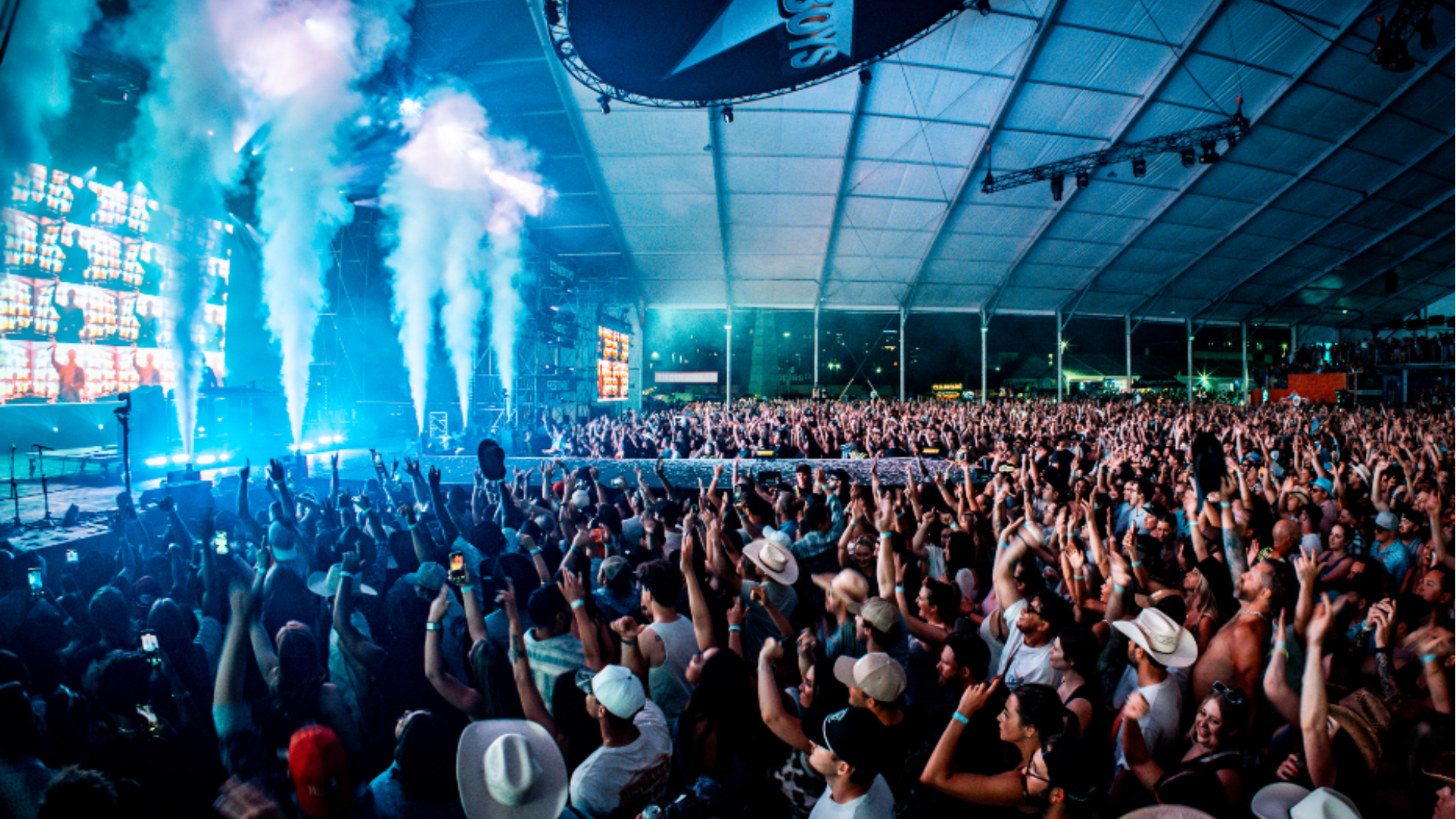 Large festival crowd raising hands as bright lights and smoke effects fill the Cowboys Music Festival tent.