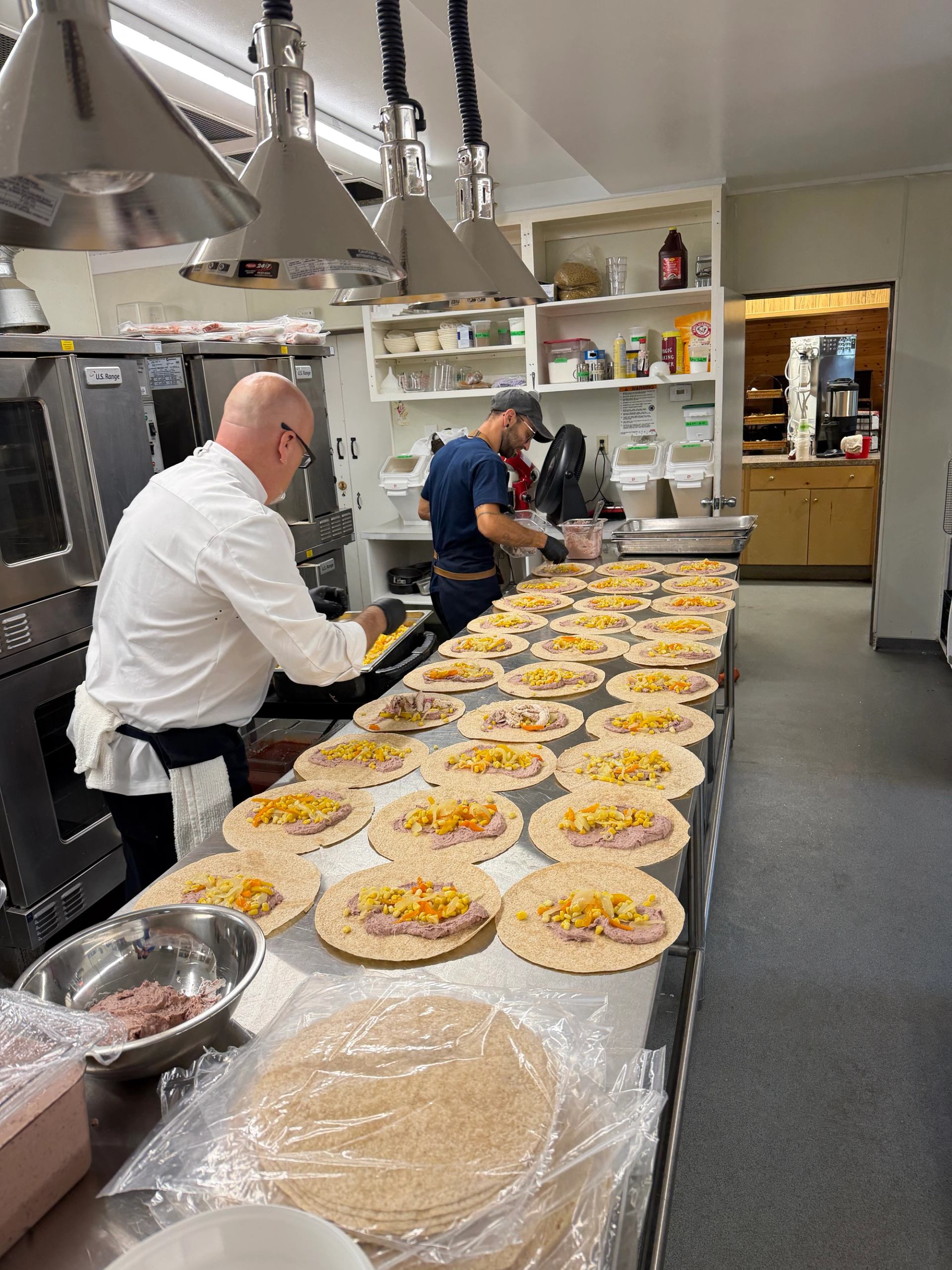 Commercial kitchen staff assembling tortilla wraps with bean spread and mixed vegetables on counter.
