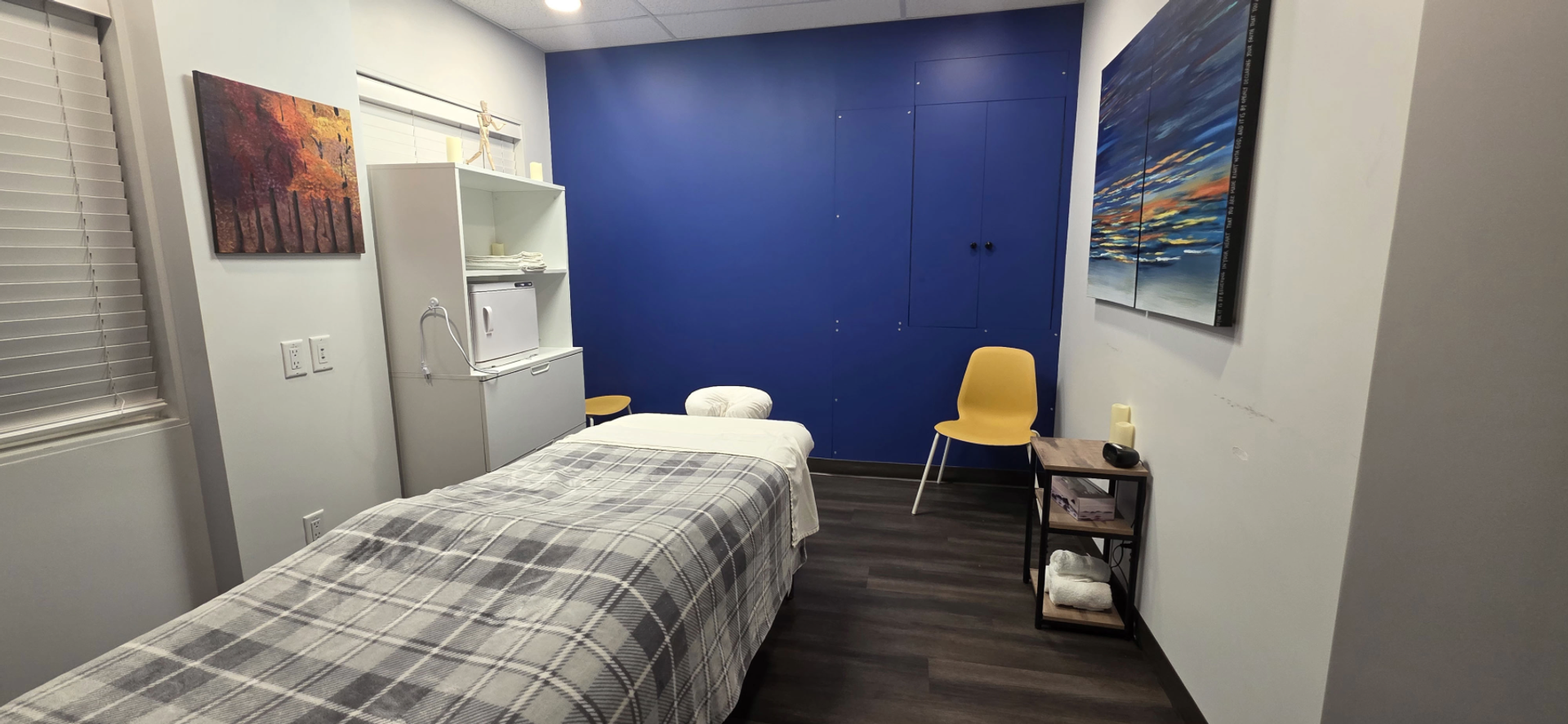 Treatment room with covered table, yellow chair, blue wall and colourful artwork.