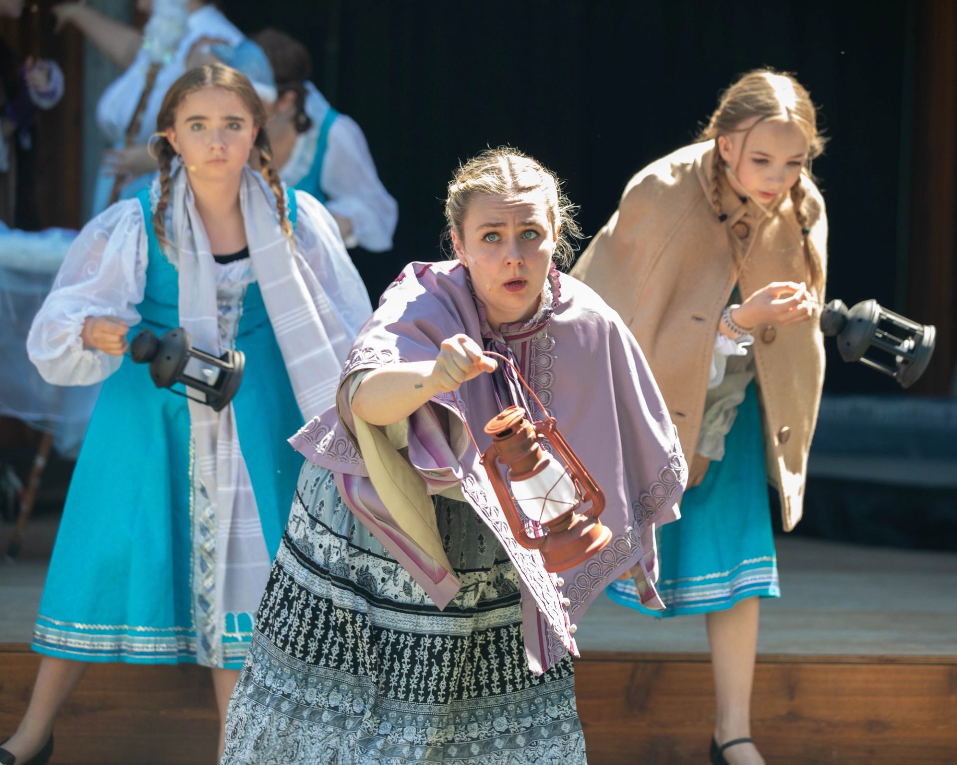 Three young actors in folk costumes hold lanterns and react with urgency during an outdoor stage performance.