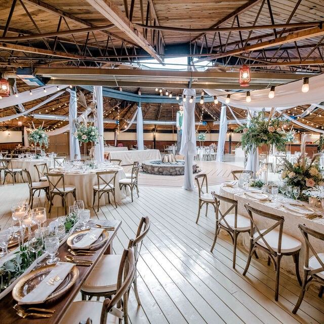 Rustic indoor wedding reception with round tables, wooden chairs, greenery, and soft string lighting.