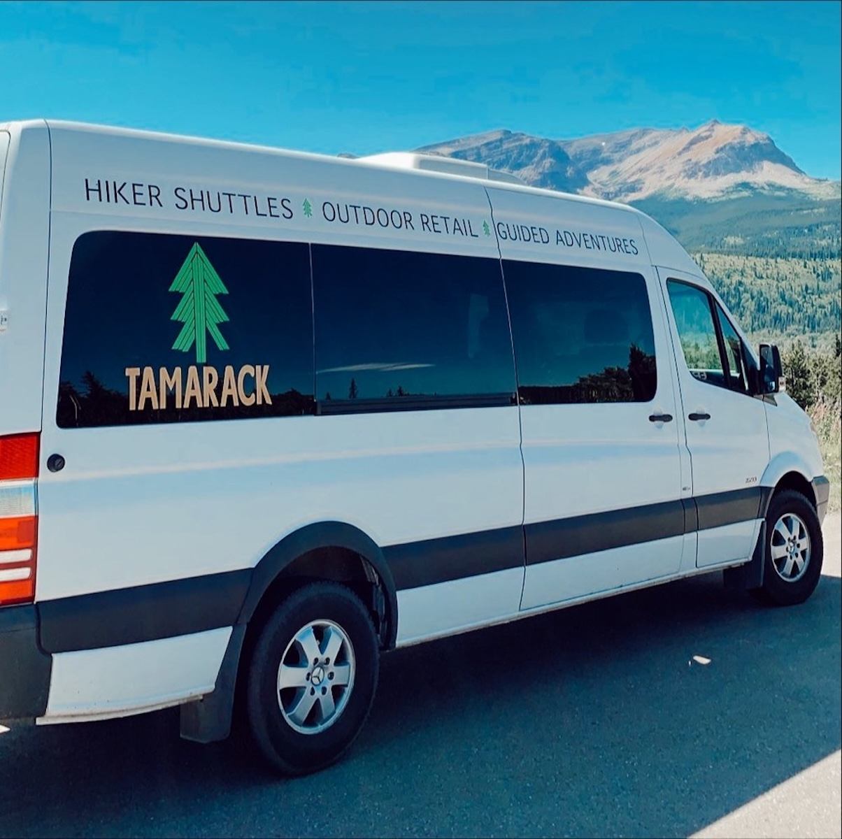 White Tamarack hiker shuttle parked on a mountain road with peaks in the background.