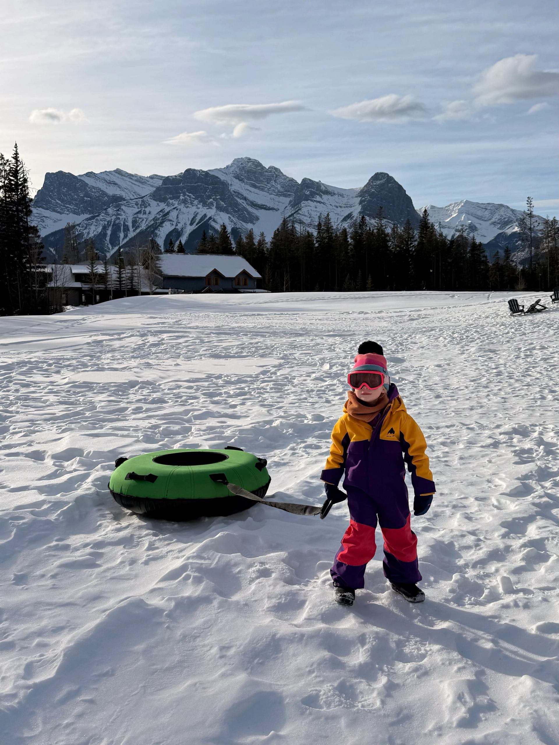 Child in a colorful snowsuit standing on a snowy field with a green tube and mountain peaks at Silvertip Resort.