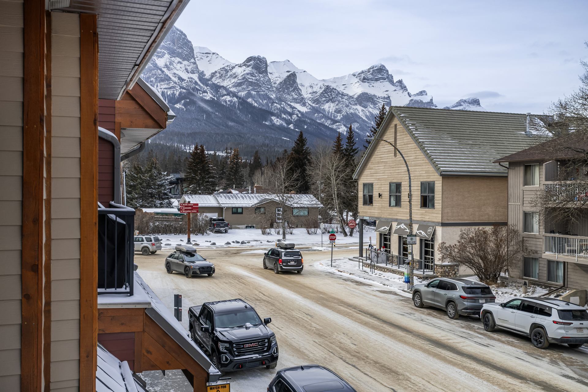 Snowy small-town street with parked vehicles, low buildings and distant rugged mountains.