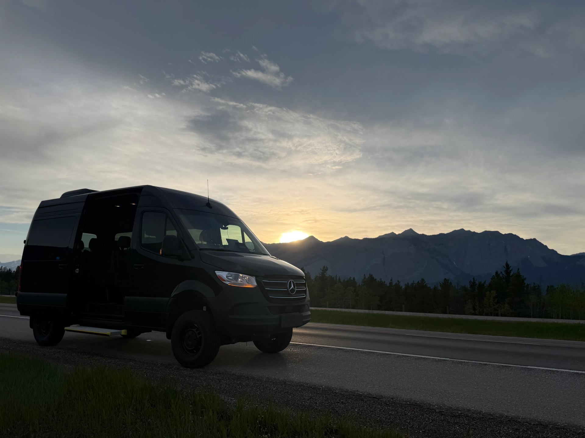 Black van parked on roadside with mountains and low sun behind distant treeline.