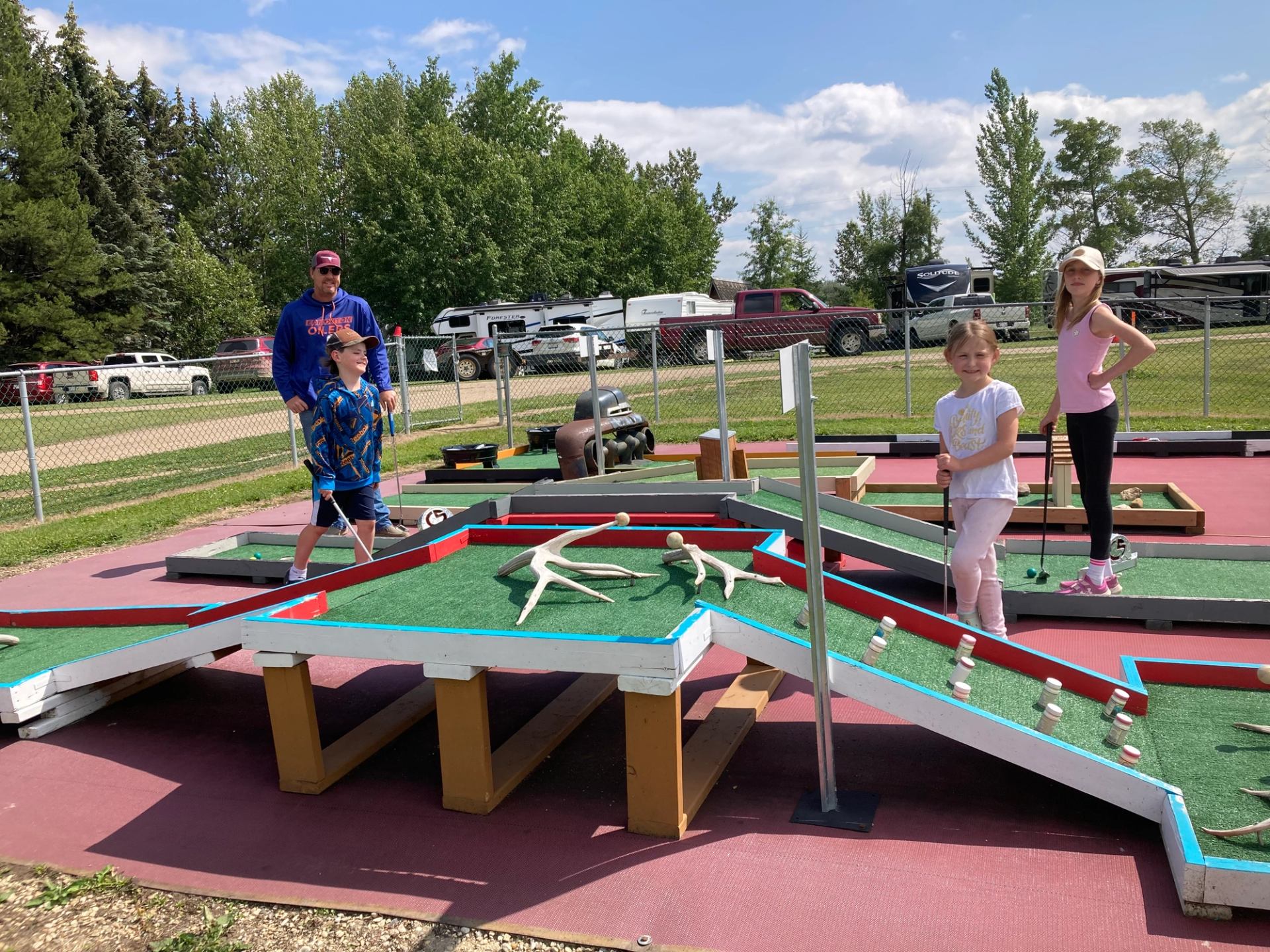 Children and adult playing mini golf on outdoor course with ramps and antler obstacles.