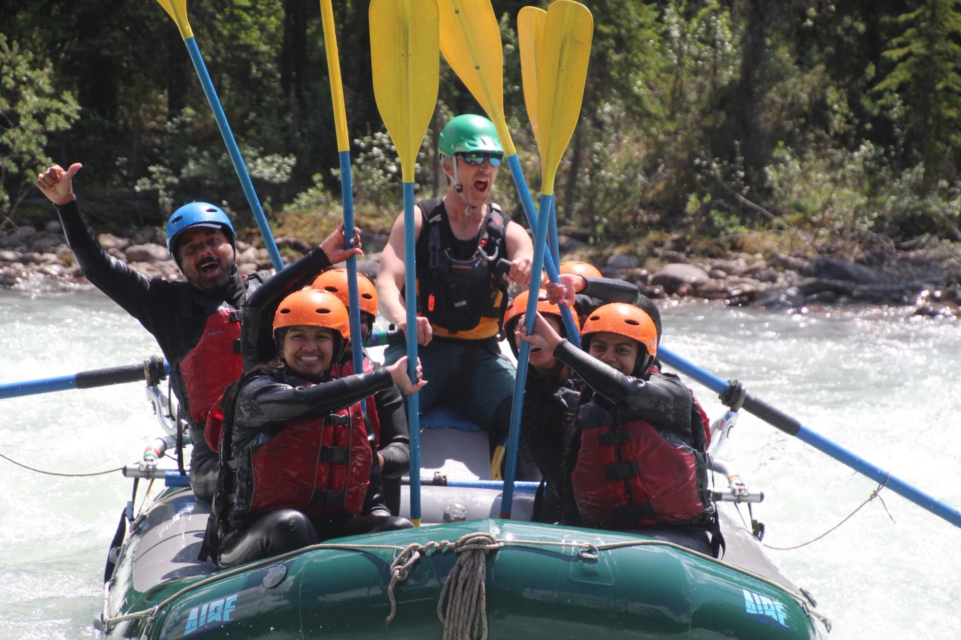 Rafting group raising paddles and cheering while floating through bright, fast-moving water.