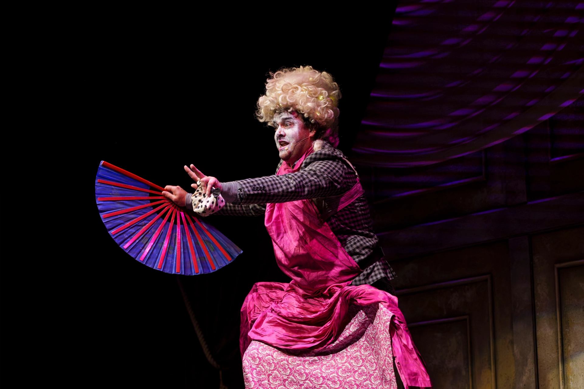 Stage performer in pink costume and wig holds colourful folding fan mid-gesture.
