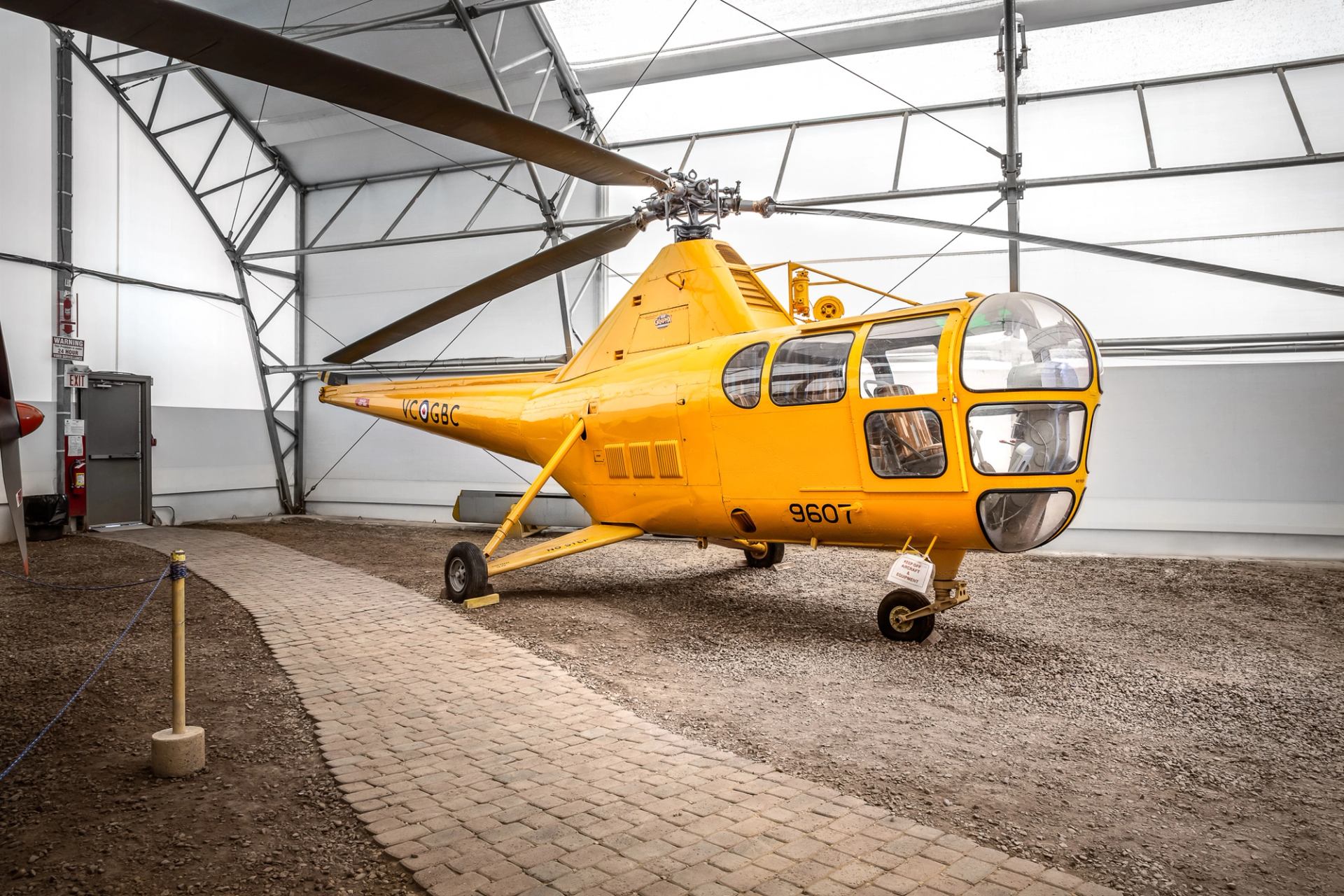 Yellow helicopter inside Hangar Flight Museum with rotor blades overhead and landing skids on floor.