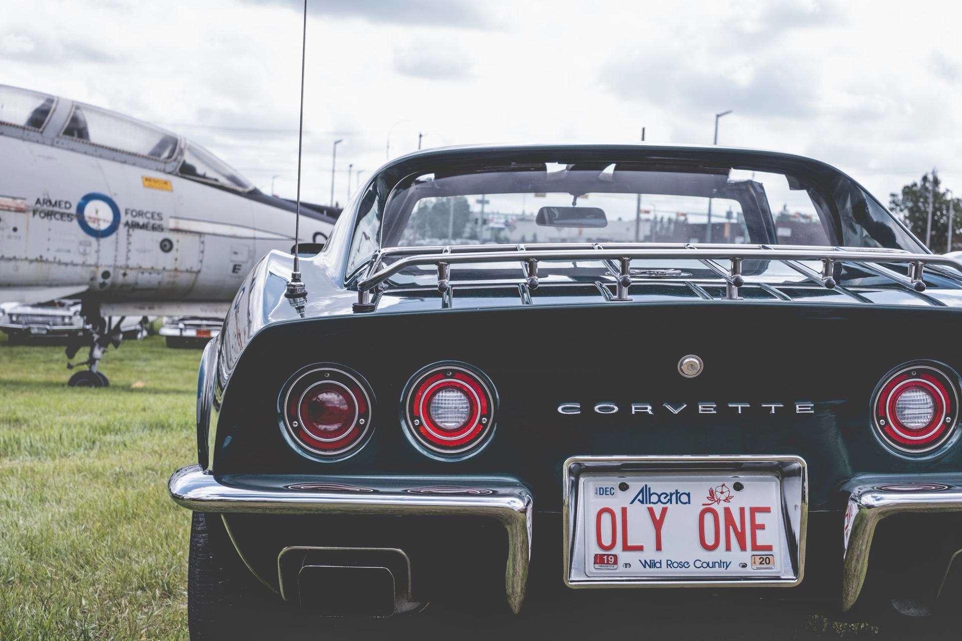 Rear view of black Corvette with Alberta OLY ONE plate beside parked military aircraft.