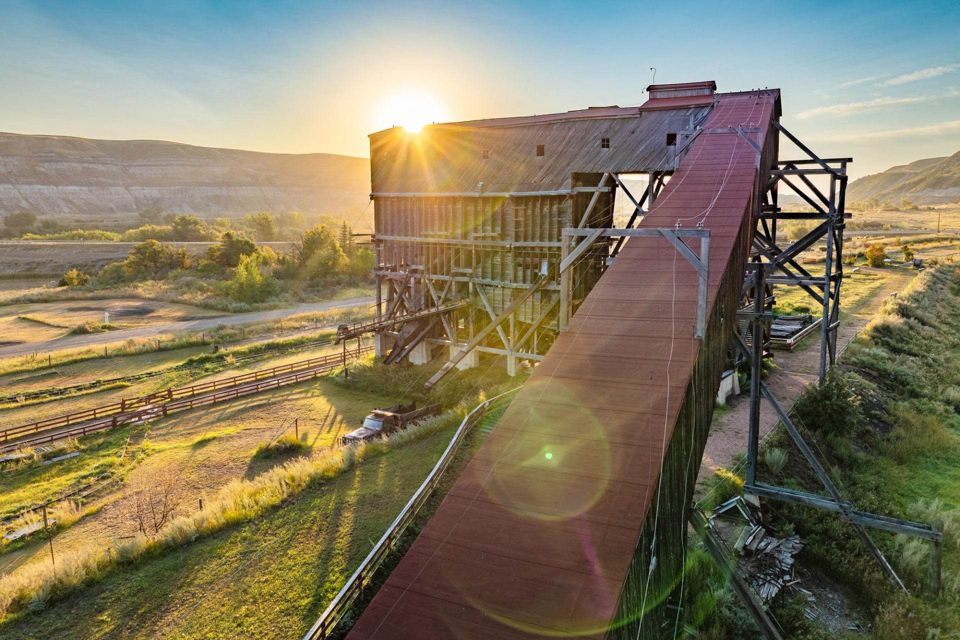 Old wooden industrial structure with long red conveyor, sun shining over rural valley.