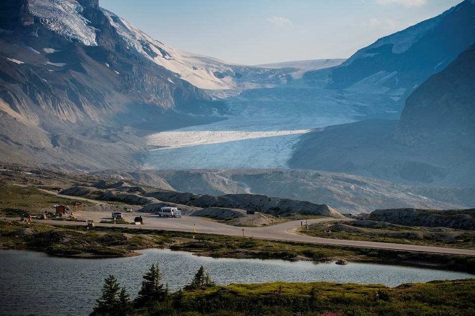 A stunning view of a glacier nestled between rugged mountains under a clear blue sky, with a winding road and parked vehicles in the foreground.