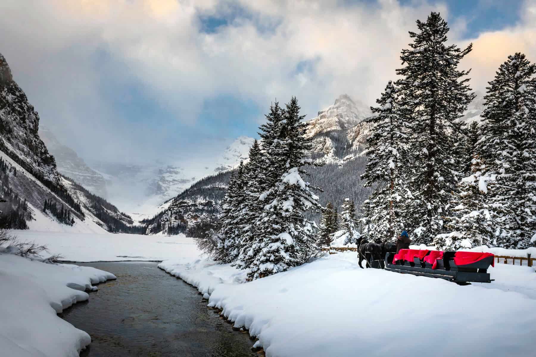 Horse-drawn sleigh beside snowy river in pine-filled mountain valley.