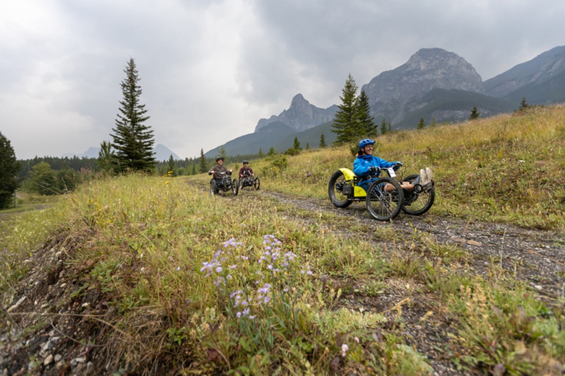 Riders on adaptive mountain bikes follow a gravel trail through grassy foothills and mountains.