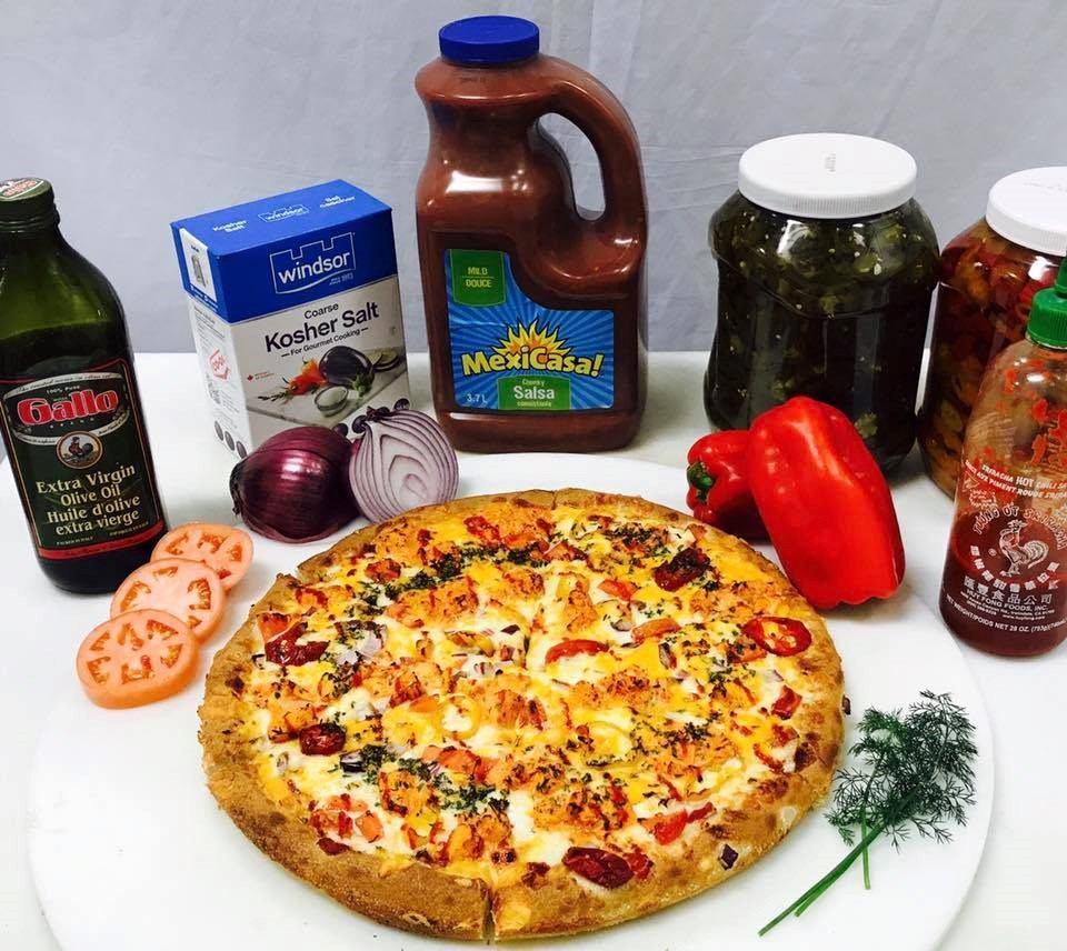 Cheese pizza on white plate with bottles, jar, sliced tomatoes and green pepper beside it.