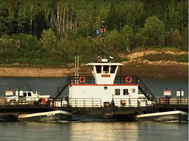 Cable ferry carrying vehicles across a river, backed by a forested hillside under clear daylight.