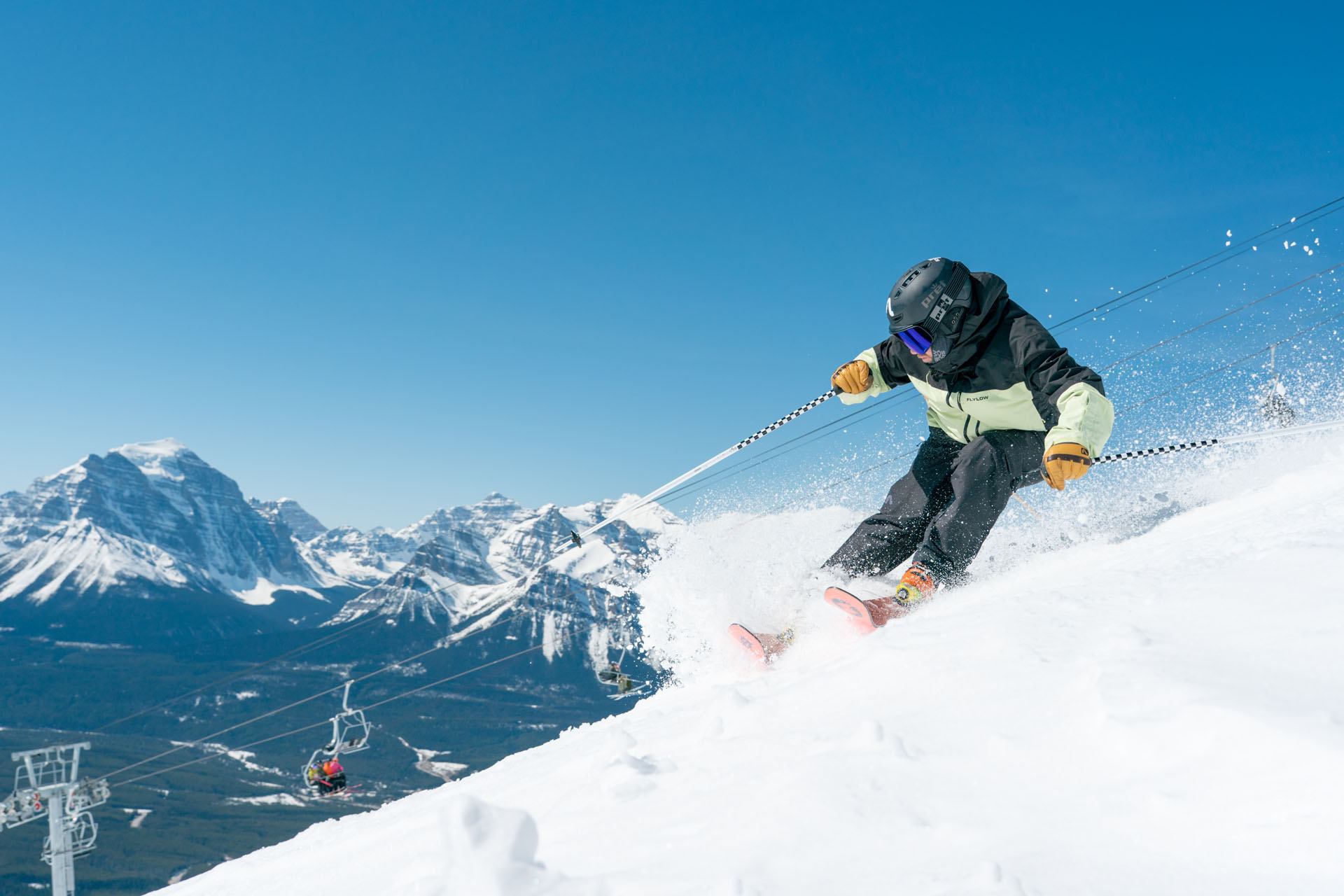 Skier carves through fresh snow on mountain slope with chairlift and peaks behind.