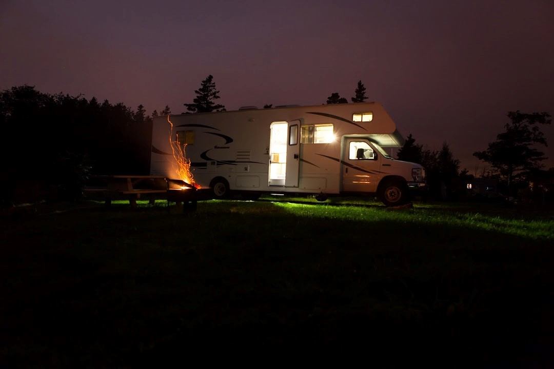 RV with lights on parked near a campfire at night in a wooded area.