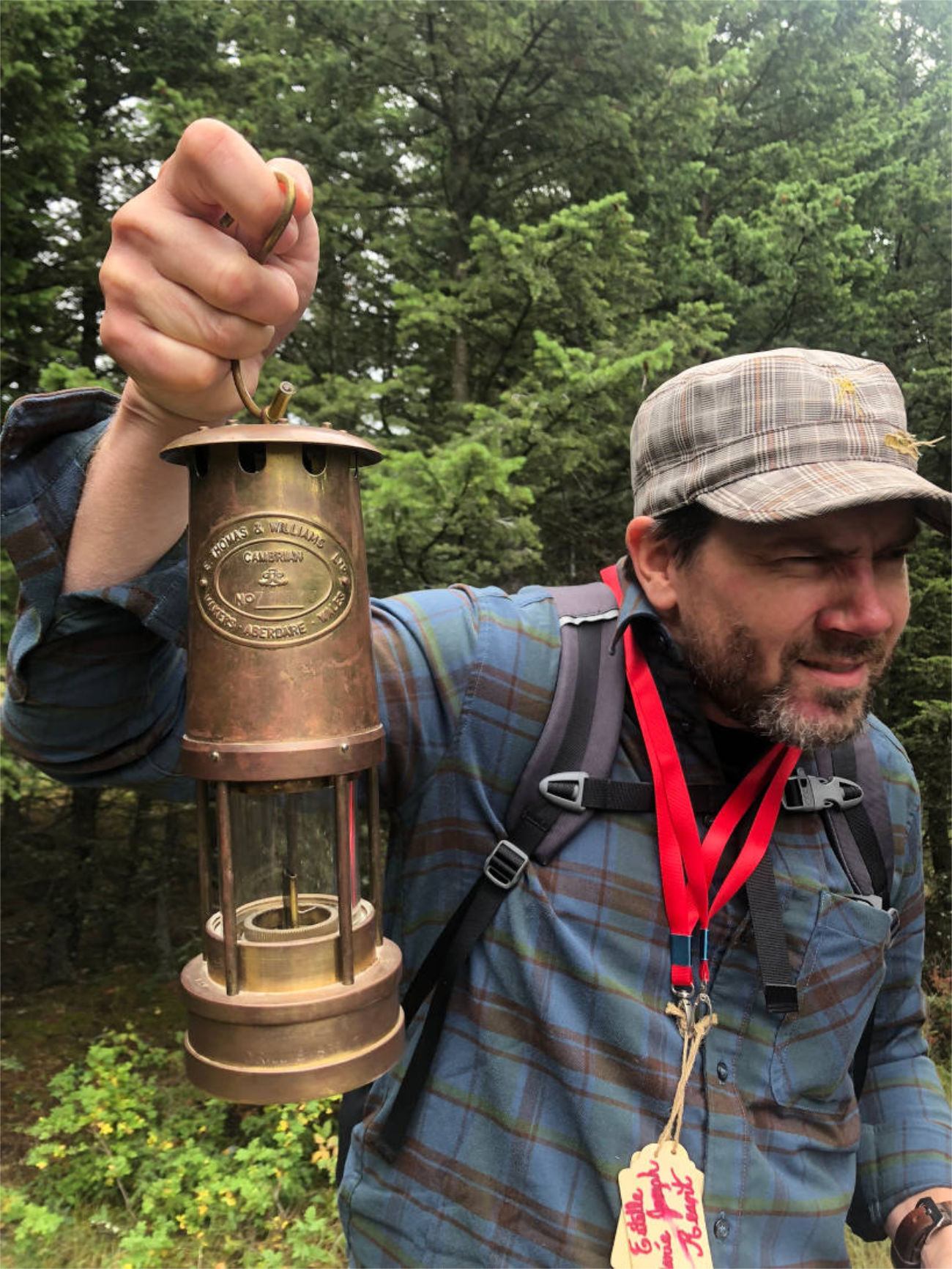 A man in a plaid shirt and cap holds up an antique brass miner's lamp in a forest.
