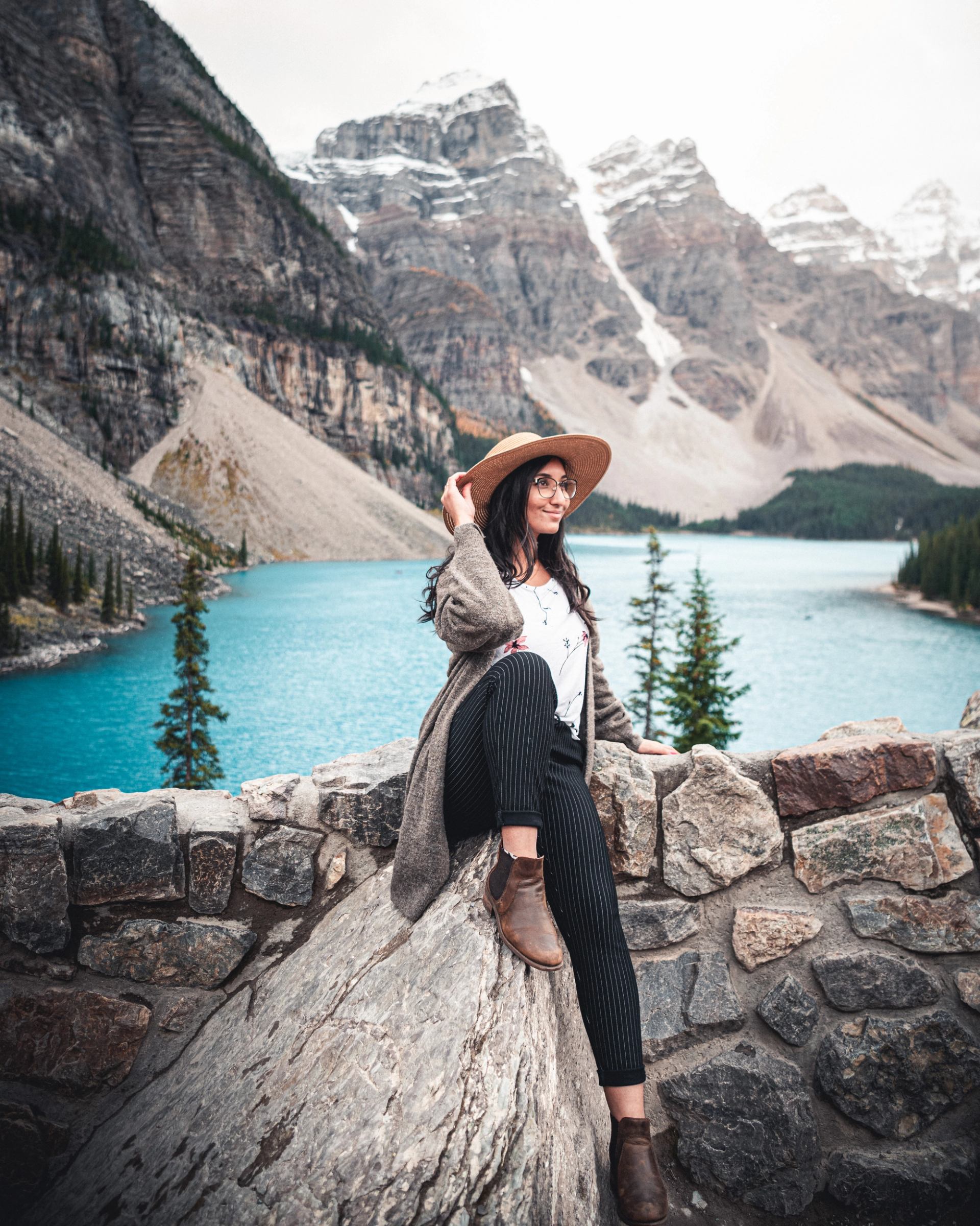 Person sitting on rock wall by turquoise mountain lake with snowy peaks.