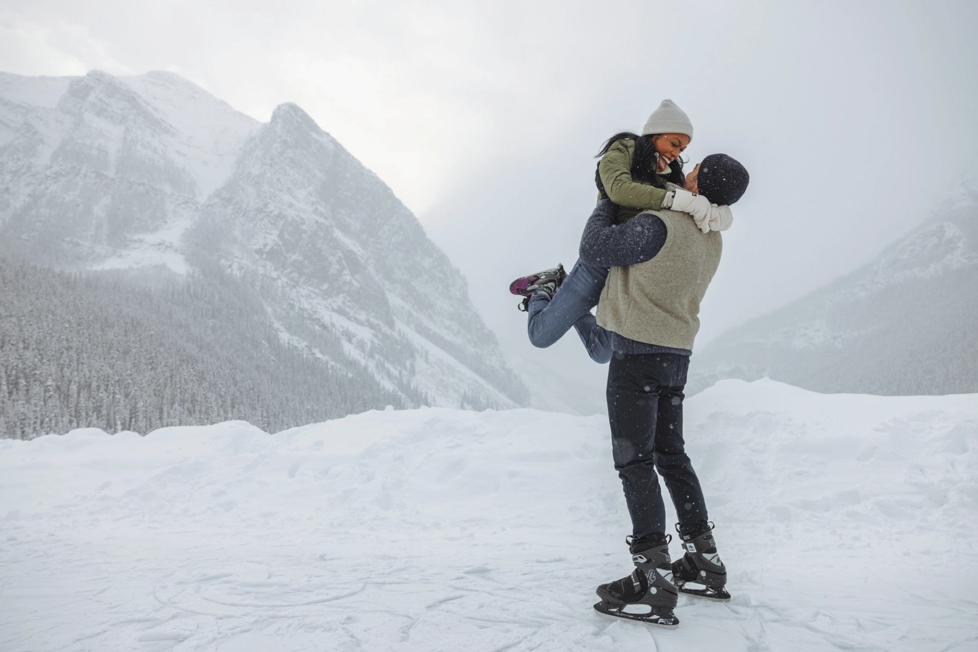 Two people in winter clothing ice skate on snowy lake with mountains behind.