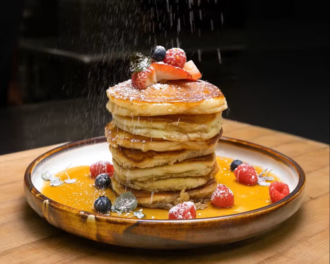 A tall stack of pancakes is topped with berries as powdered sugar falls onto the plate.