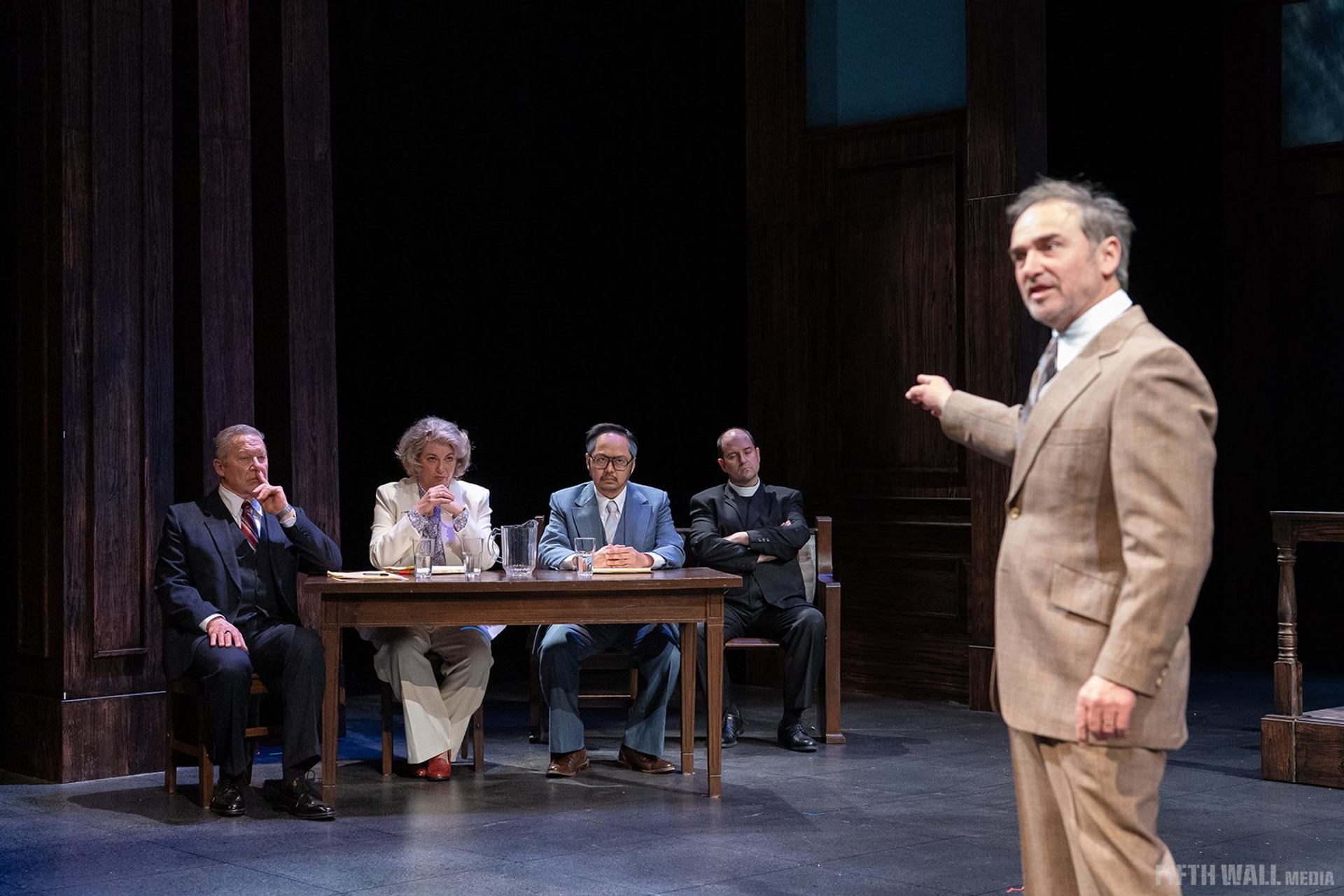 Stage actors in formal suits sit at a wooden table as another gestures nearby.