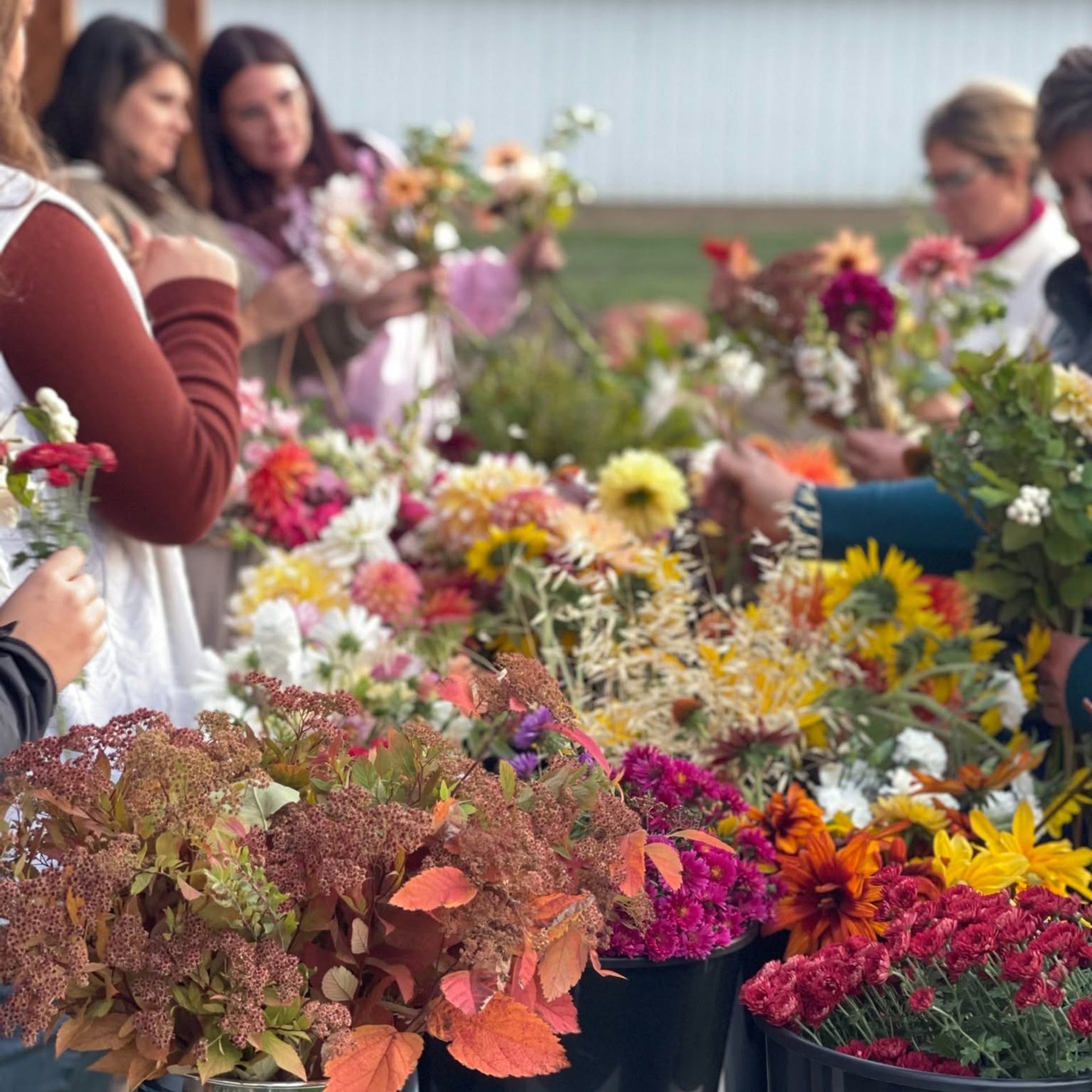 Buckets of colourful cut flowers on a table with people arranging bouquets behind.