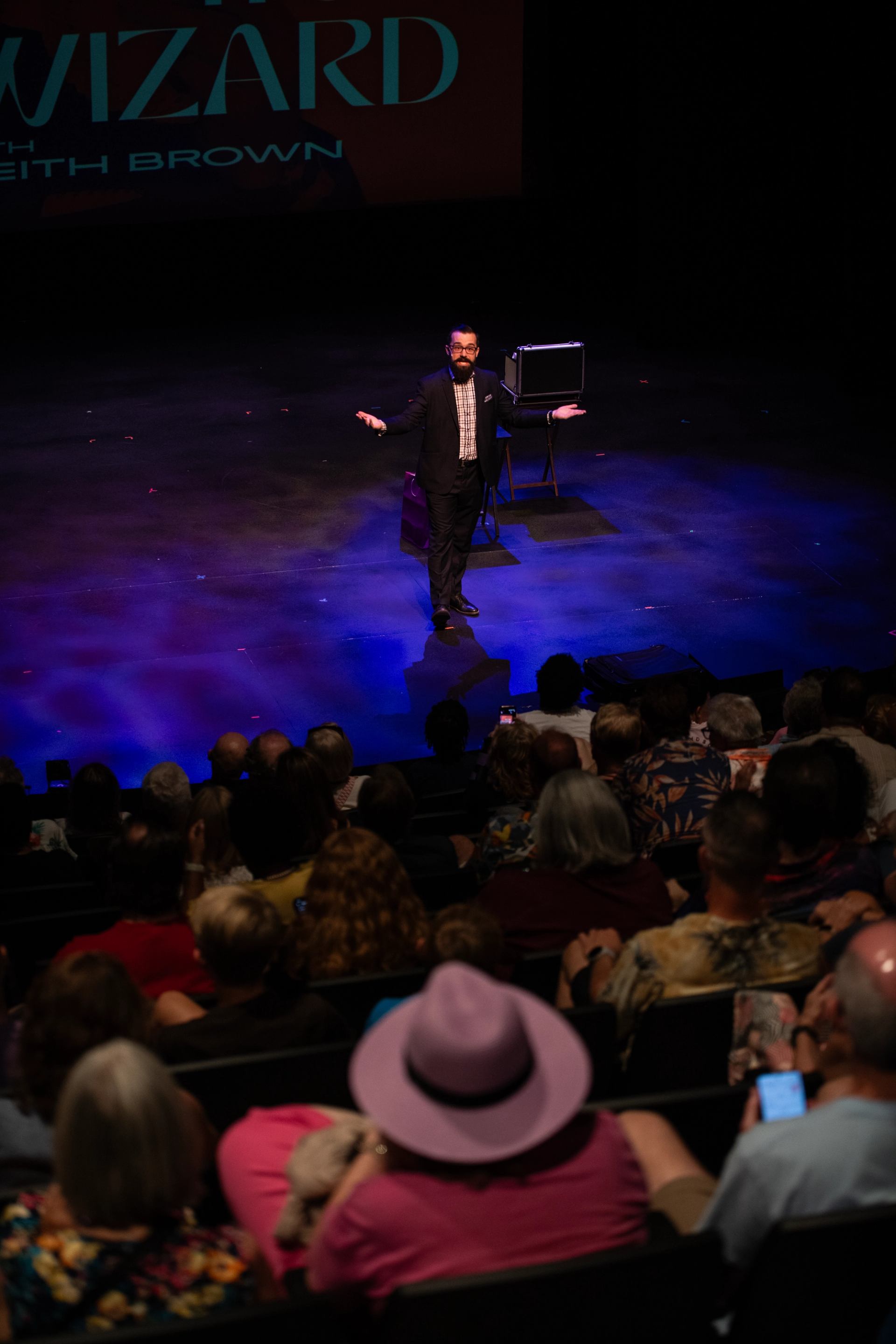 Stage performer in suit addresses seated theatre audience under blue and purple lighting.