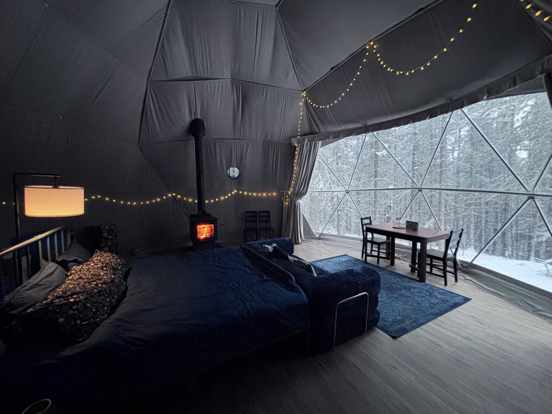 Geodesic dome interior with bed, wood stove, string lights and snowy forest view.