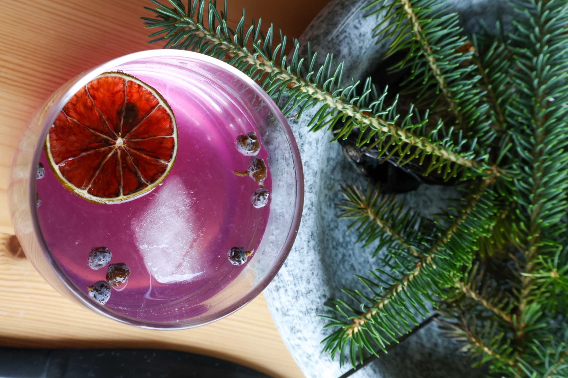 Purple cocktail with citrus slice and juniper berries beside fresh pine sprigs.