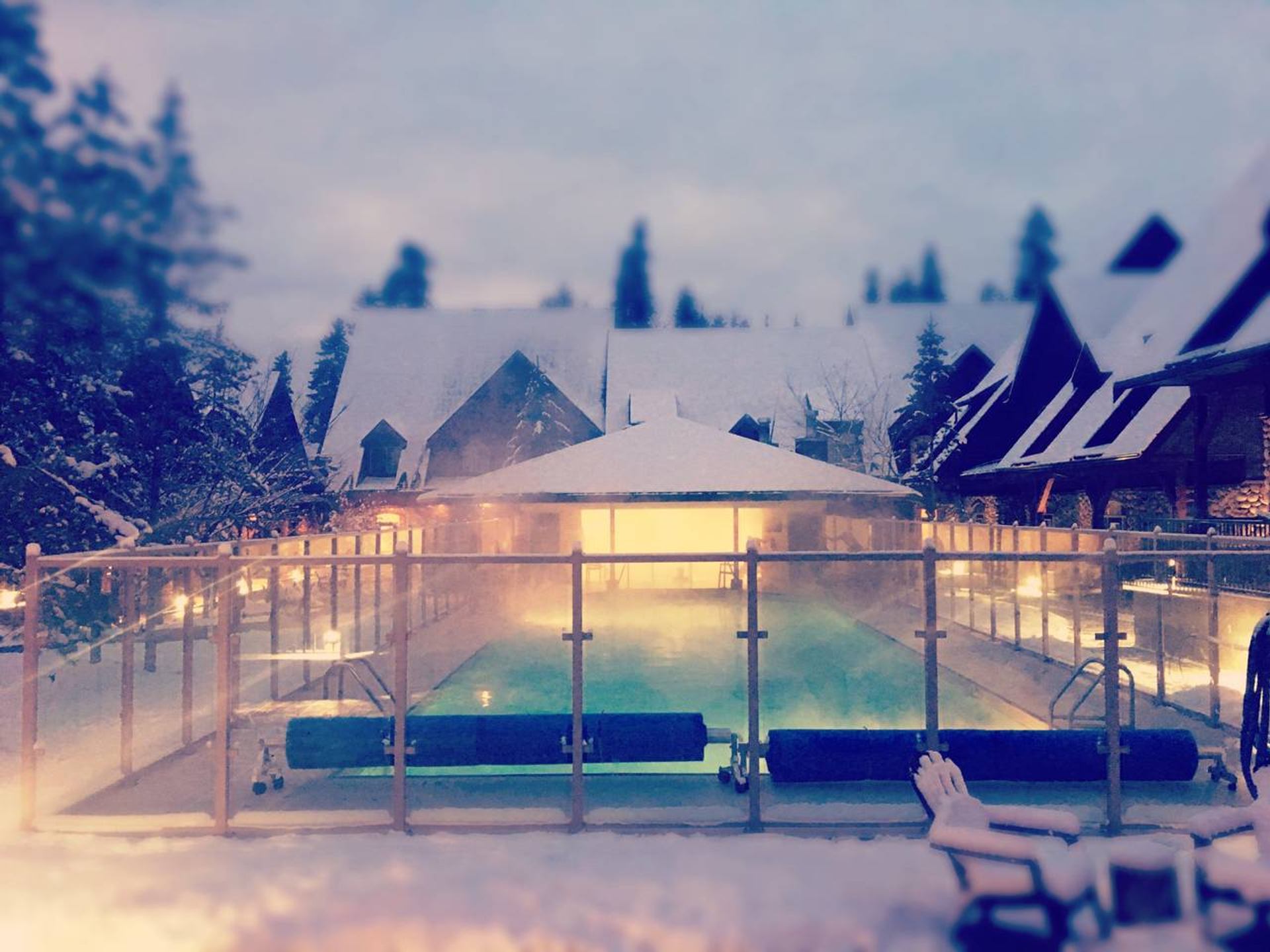 Snow-covered outdoor pool glows behind glass fencing, surrounded by chalet-style buildings.