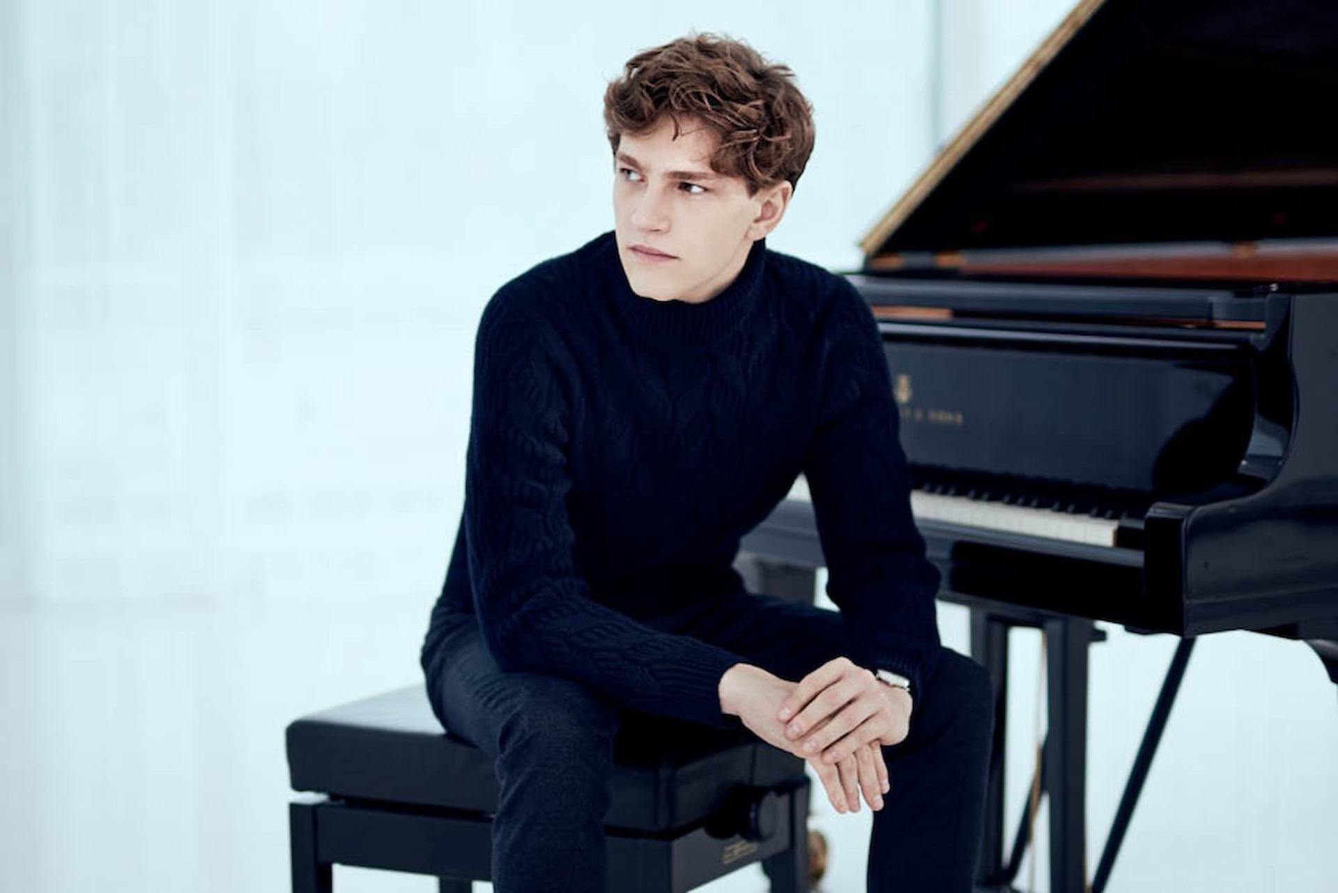 Person in dark sweater sits on piano bench beside grand piano in bright room.