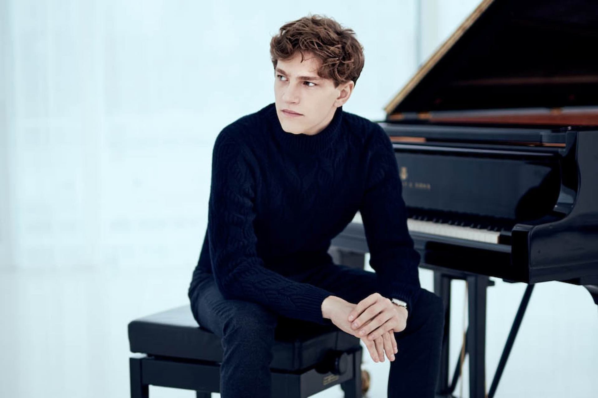 Person in dark sweater sits on piano bench beside grand piano in bright room.