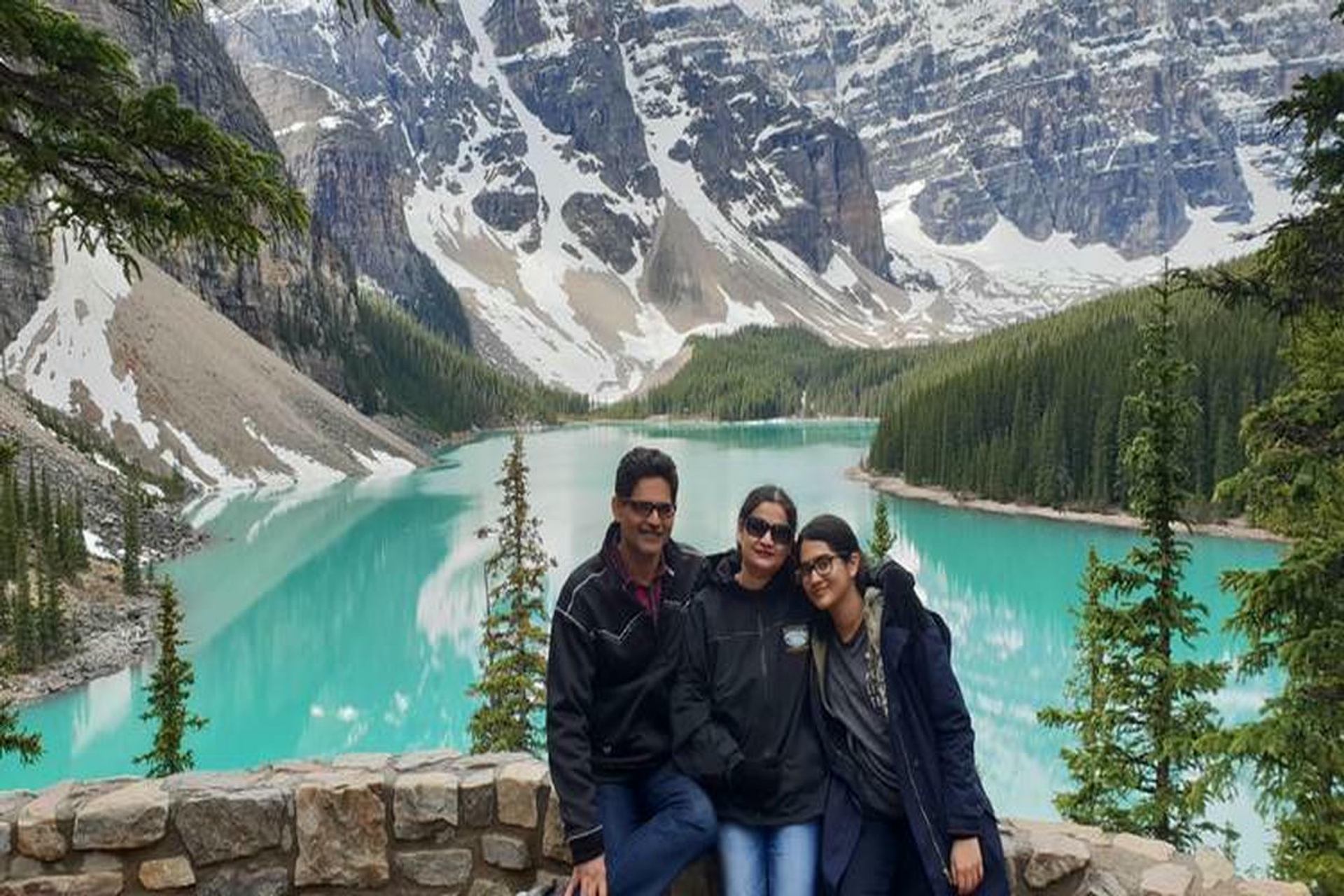 Three people stand by turquoise mountain lake with snow-capped peaks and evergreen trees.
