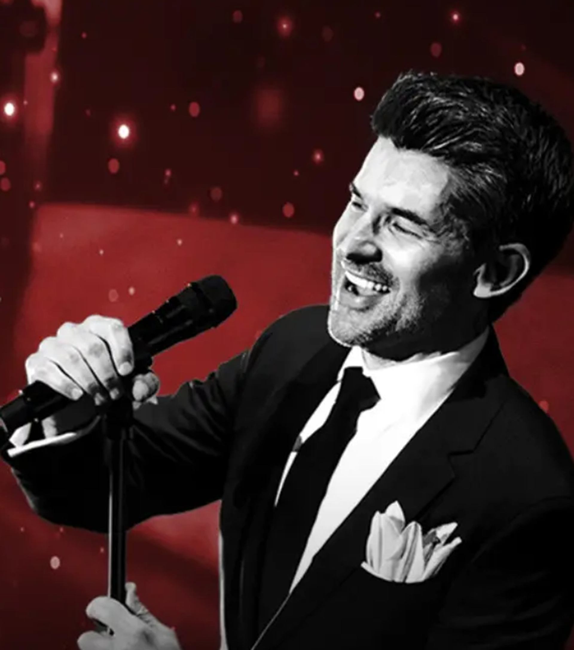 Male singer in suit holding microphone on stand against red, sparkling background.