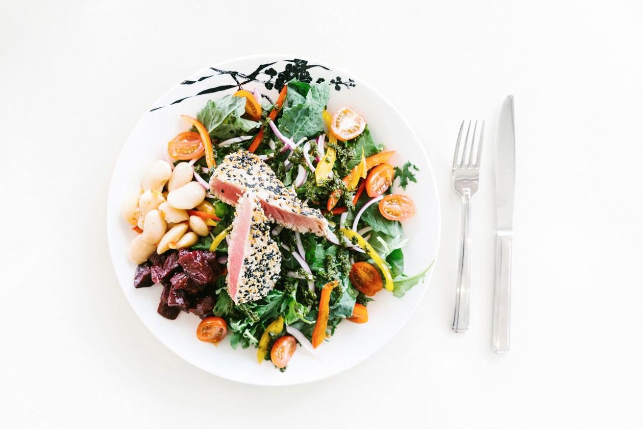 Seared sesame tuna salad with mixed greens, white beans, and beets on a white plate with a fork and knife.