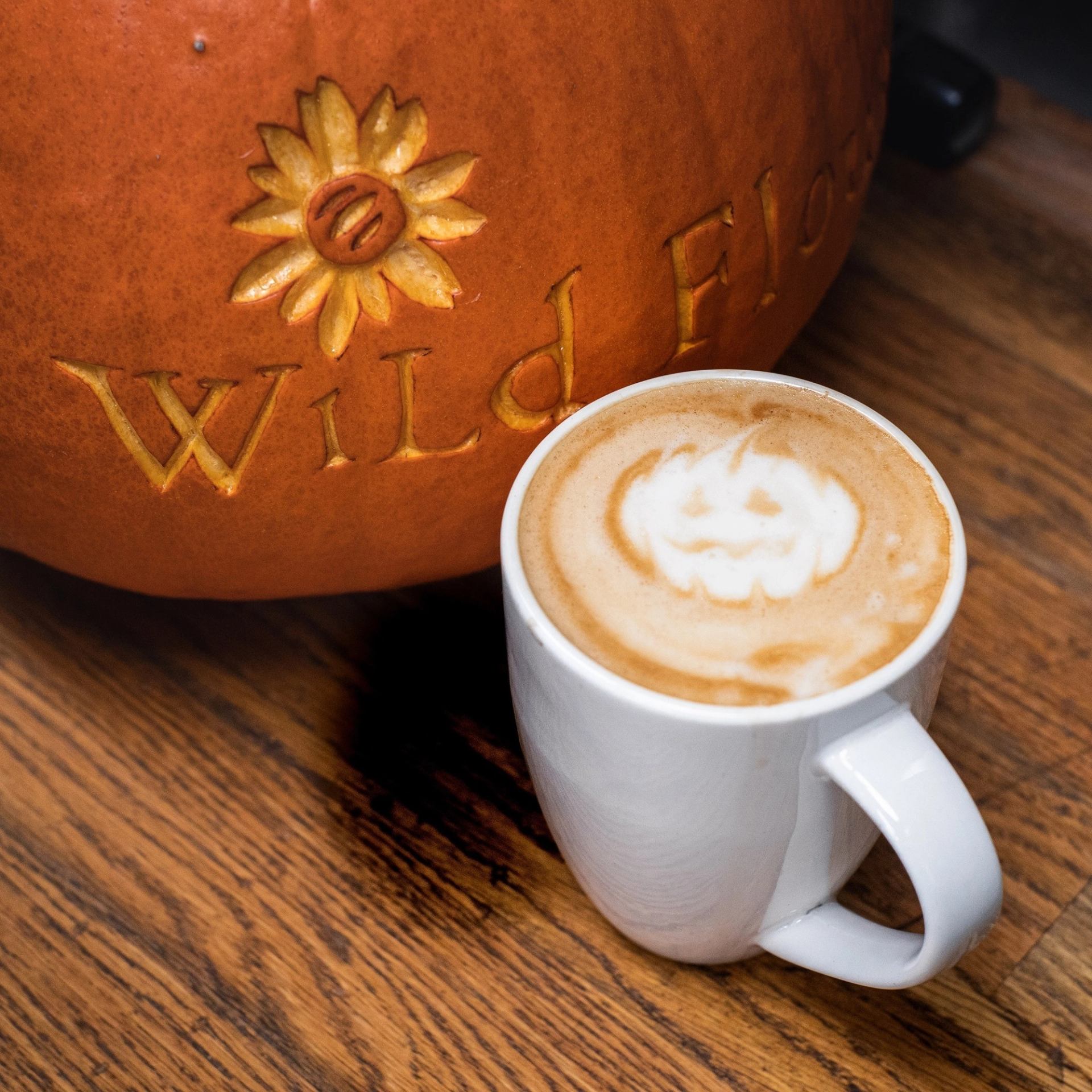 Pumpkin latte with jack-o’-lantern art beside carved Wild Flour pumpkin.