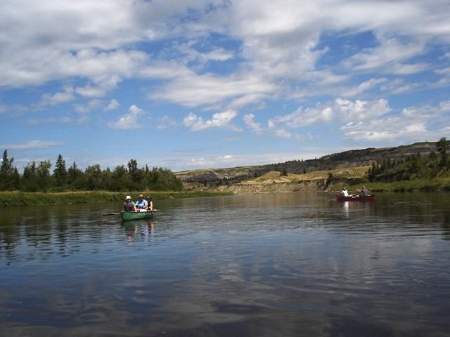 Canoes glide along a calm river with tree-lined banks and distant hills under clouds.