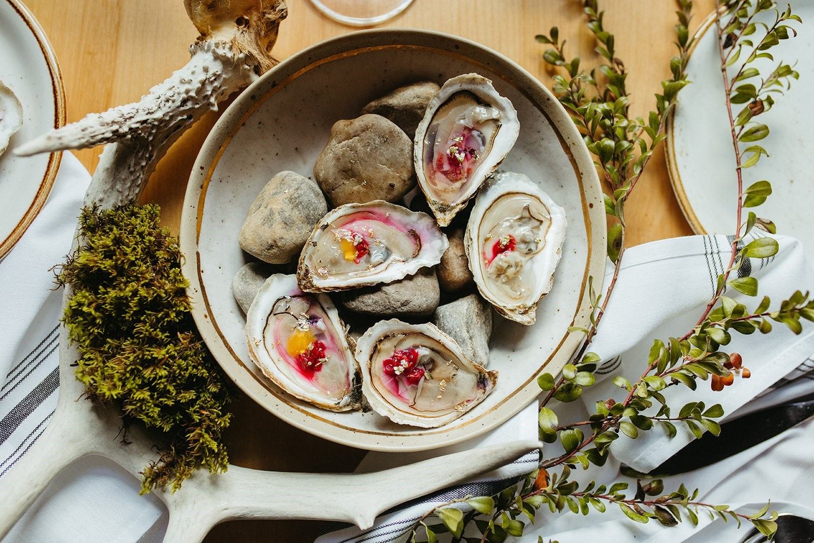 Oysters on a bed of stones with garnish, set on a wooden table with greenery around.