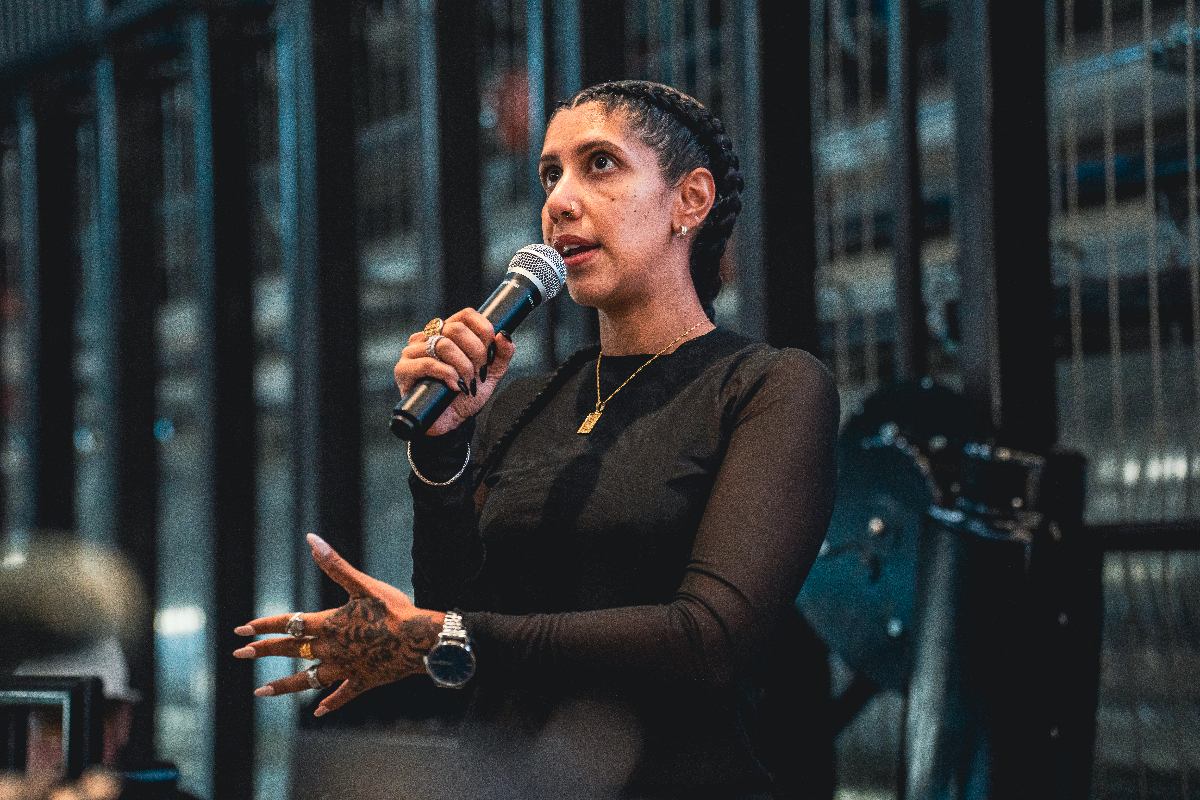A woman with braids, a black shirt and jewelry holds a microphone and gestures while delivering a speech.