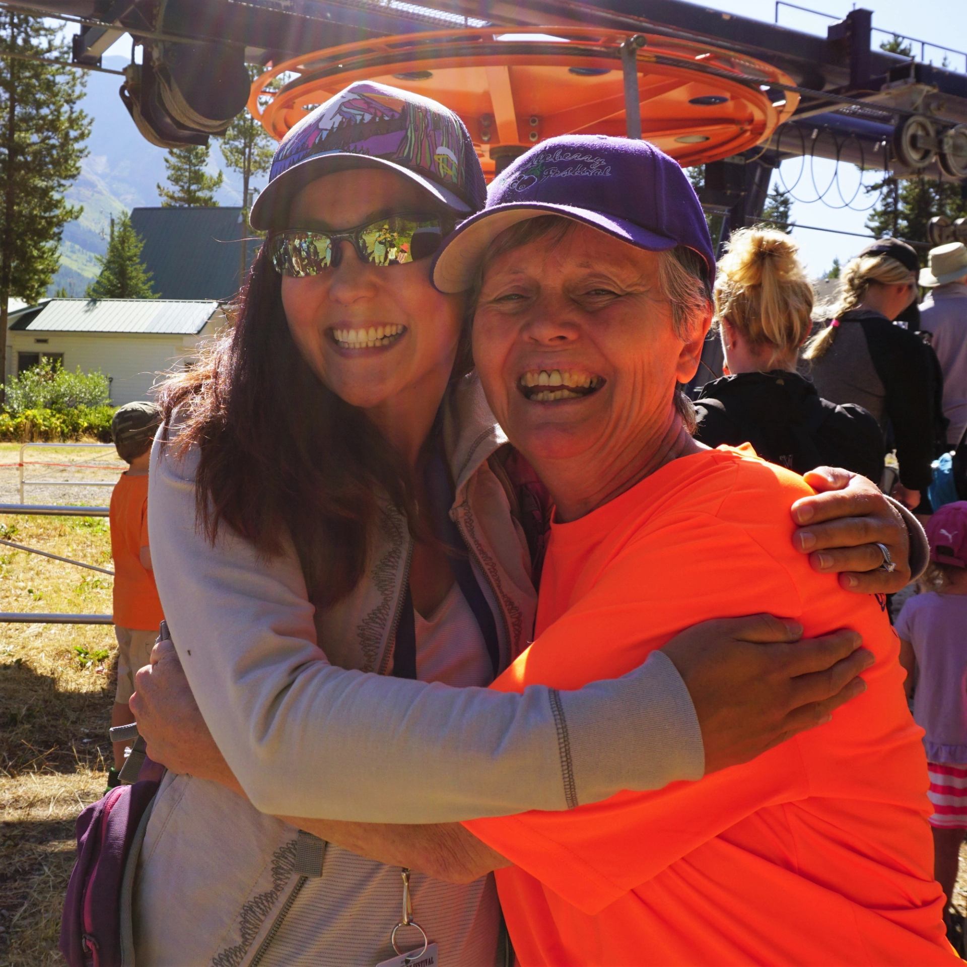 Two people wearing caps hug outdoors near a ski lift with others behind them.