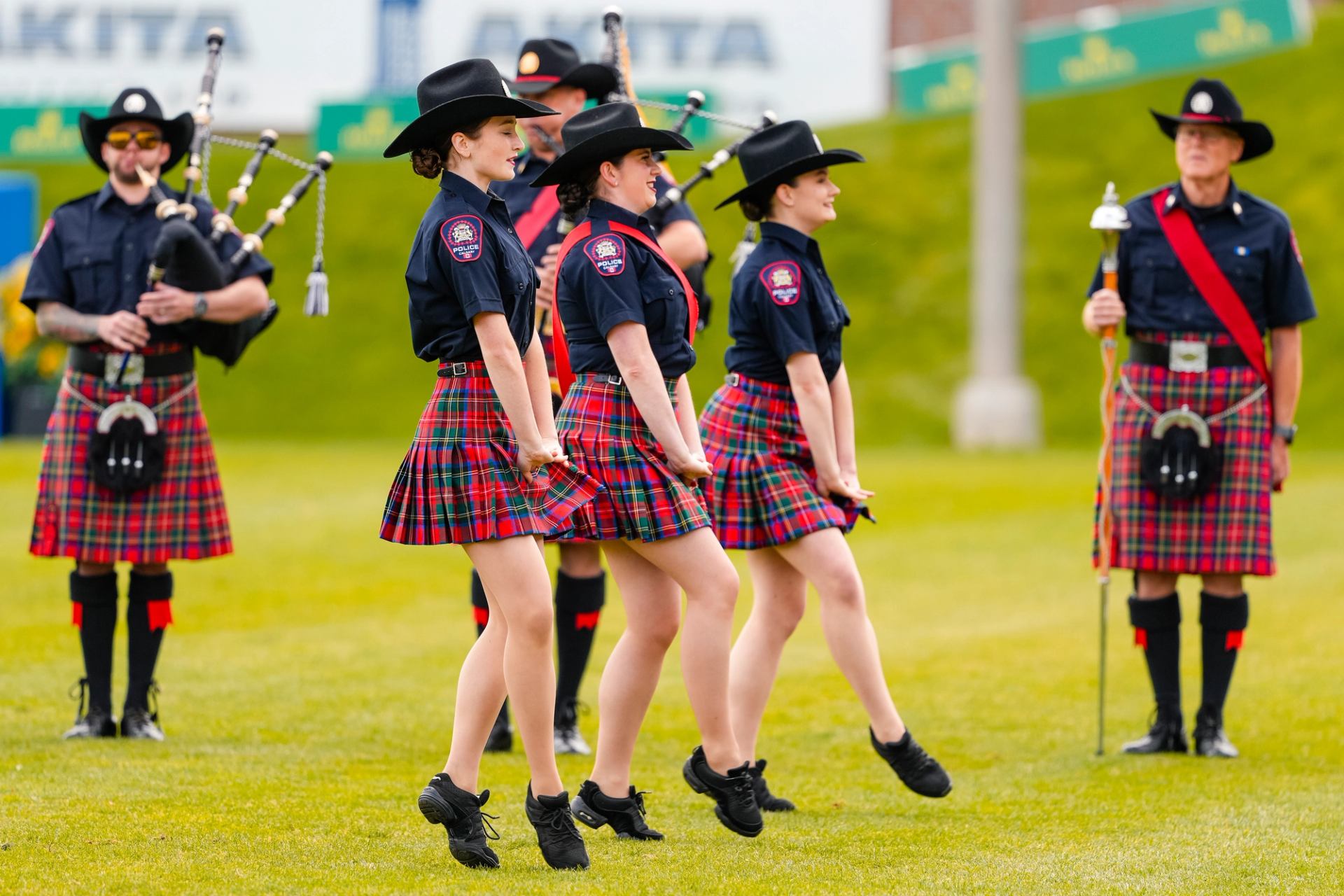 Dancers in tartan skirts and uniform tops perform while pipers stand behind them.
