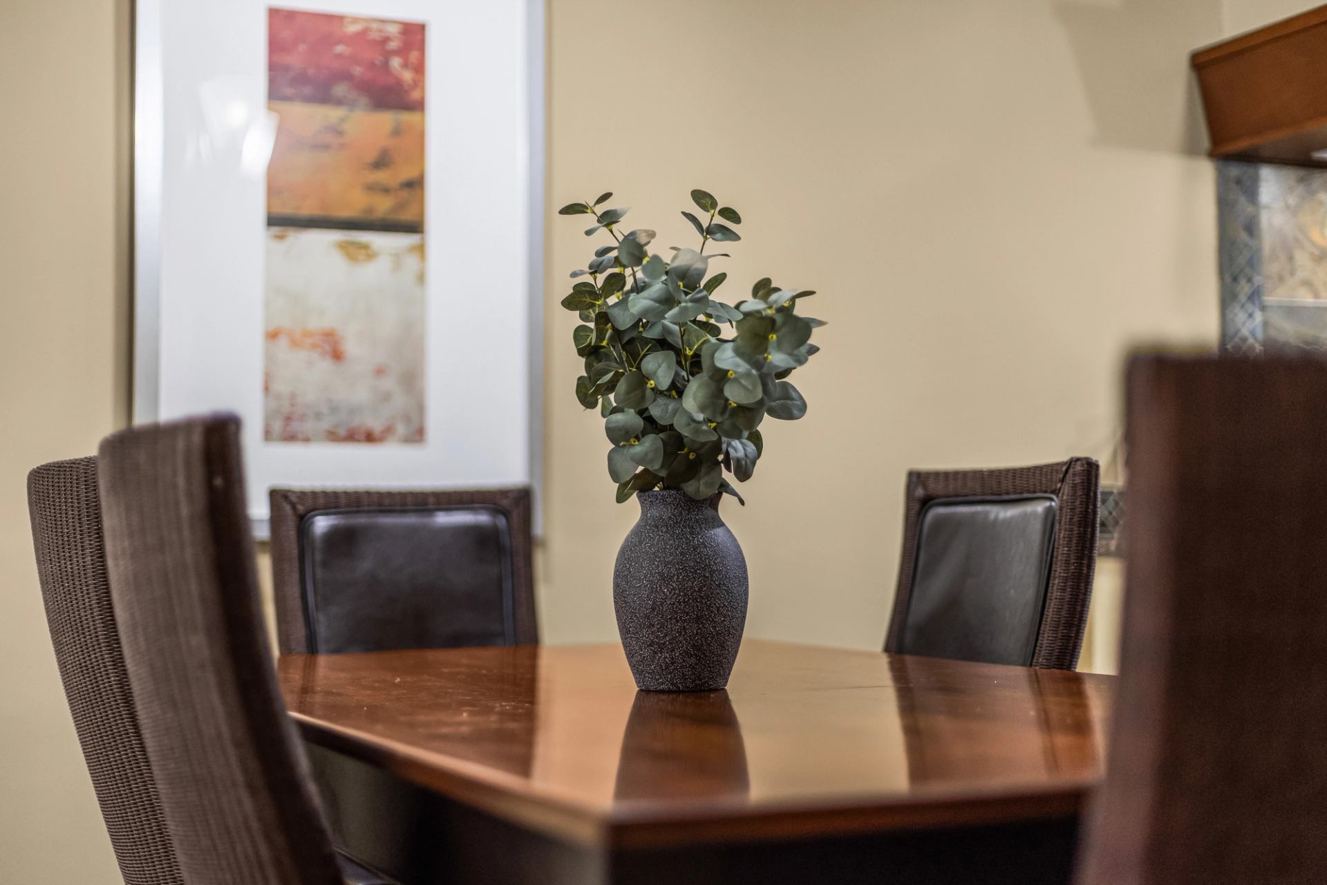 Vase of leafy branches centred on a wooden dining table surrounded by chairs.