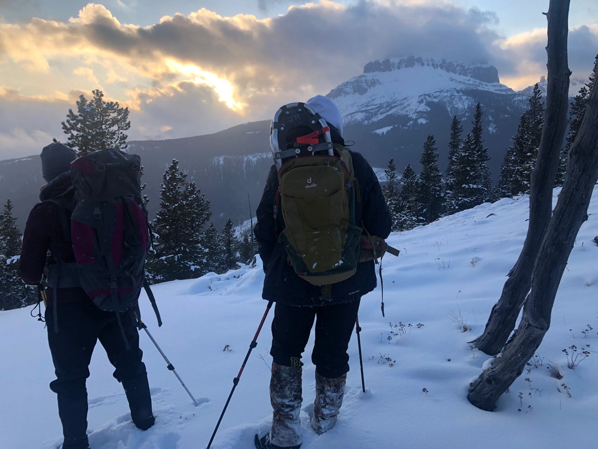 Two hikers snowshoeing at sunset on McGillivray Ridge with a rugged peak in the distance.